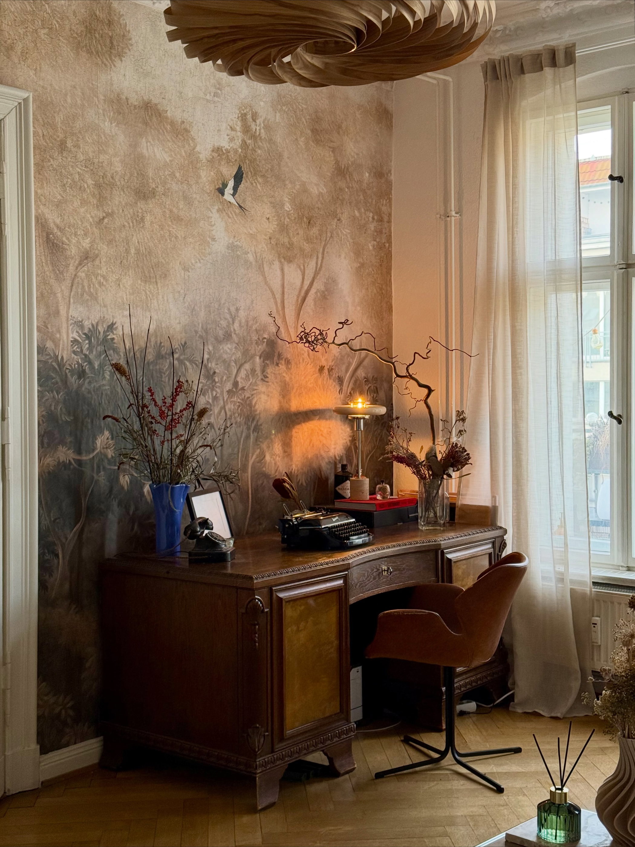 A study with a vintage wooden desk, black typewriter, and gold lamp. The wall has a forest mural with a bird. A brown chair is at the desk.