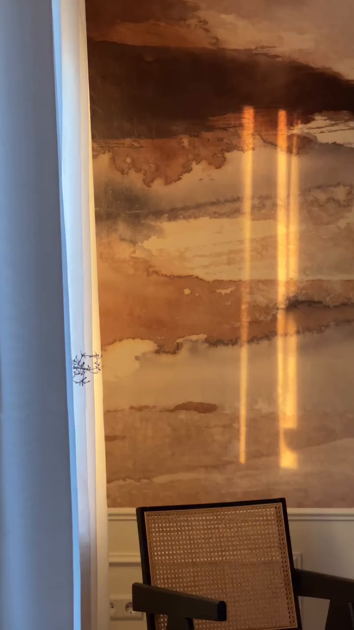 Interior with abstract wall art, sunlight streaks, and a cane-backed chair.