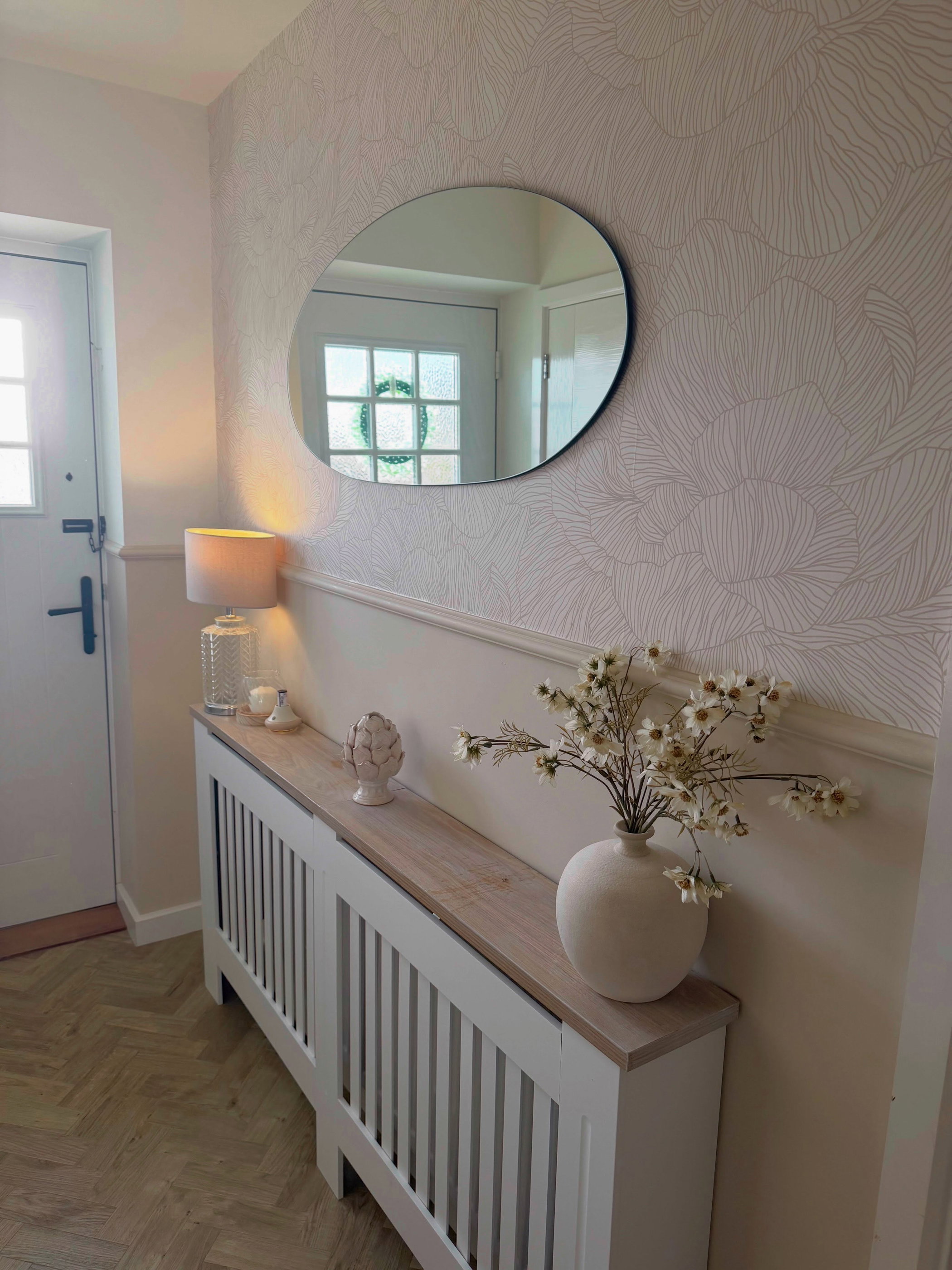White radiator cover with wood top, holding a lamp and white vase of flowers, in a bright hallway. An oval mirror reflects a front door.