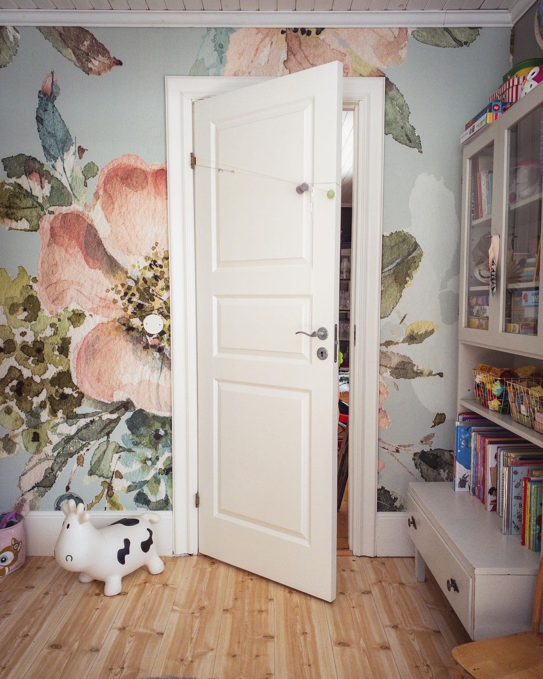 Kinderzimmer mit Blumentapete, offener weißer Tür und Kuhspielzeug auf Holzboden.