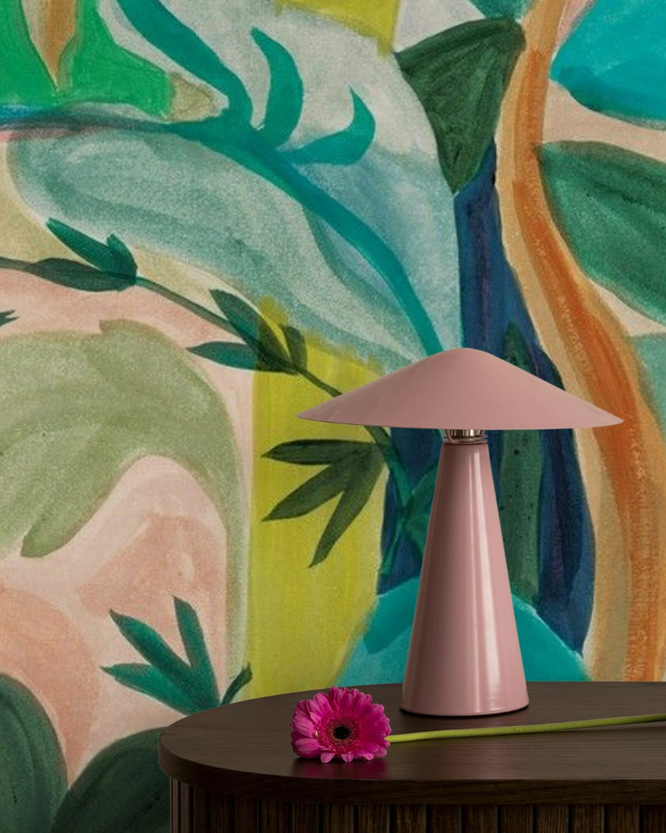 Pink table lamp with a flower on a wooden table against a colorful botanical wallpaper.