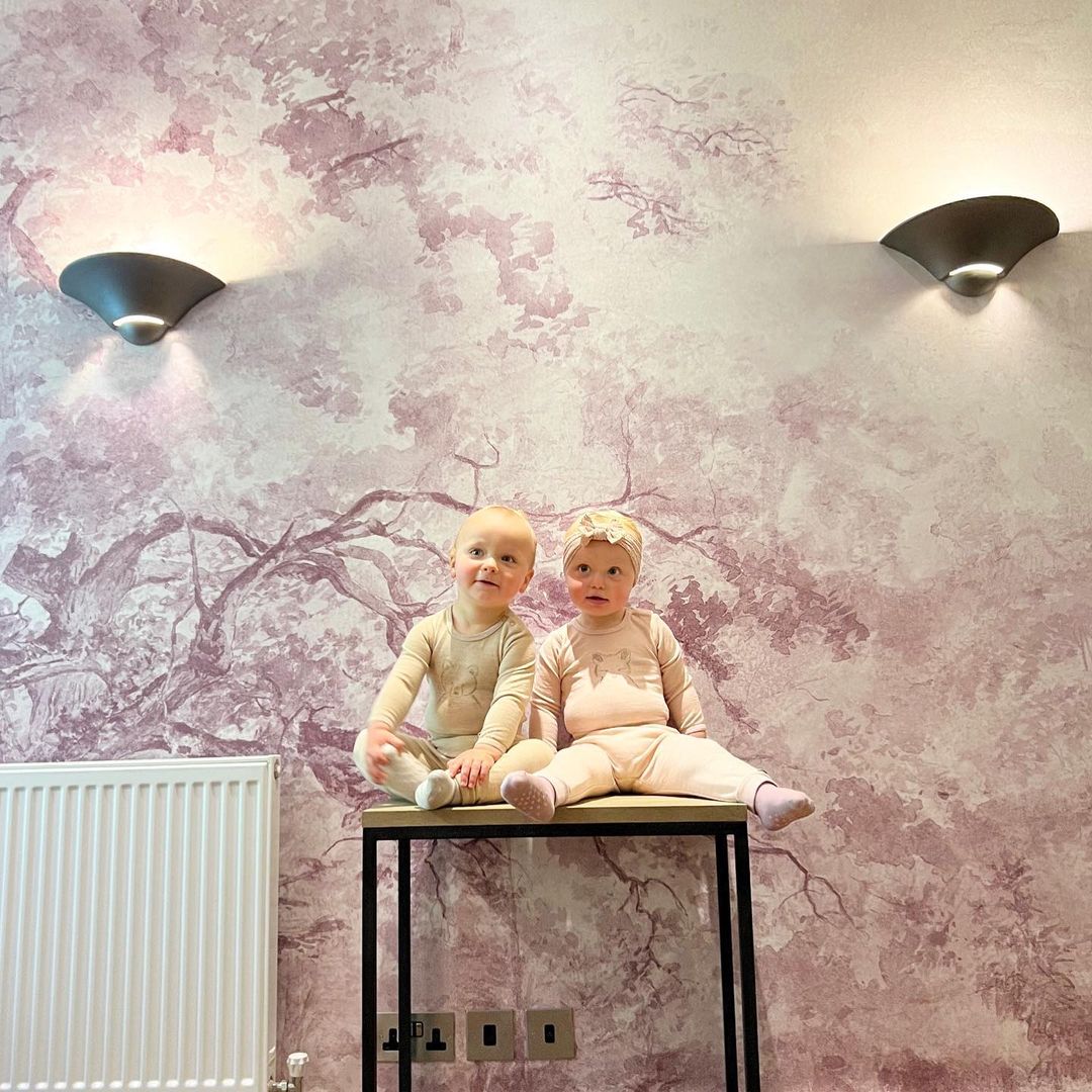 Deux bébés sont assis sur une table devant un papier peint rose sur le thème des arbres, éclairés par des appliques.