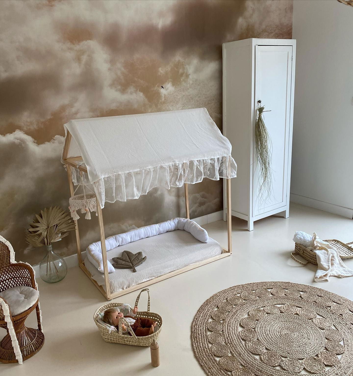 Nursery with wooden canopy bed, white linens, and cloud wallpaper. Wicker chair and rug add texture.
