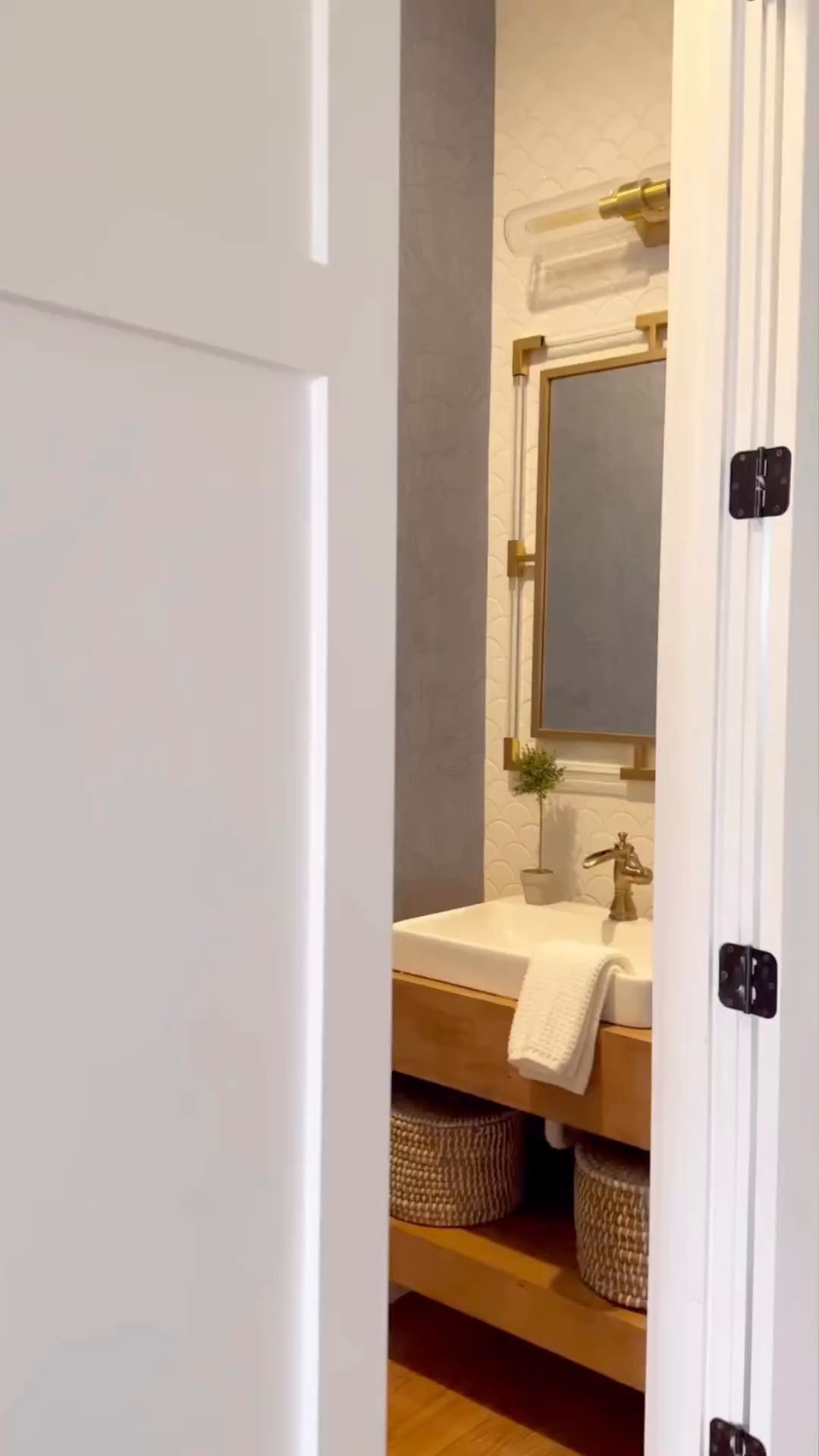Bathroom peek through doorway. Vanity with white sink, gold faucet, and mirror. Wicker baskets on shelf.