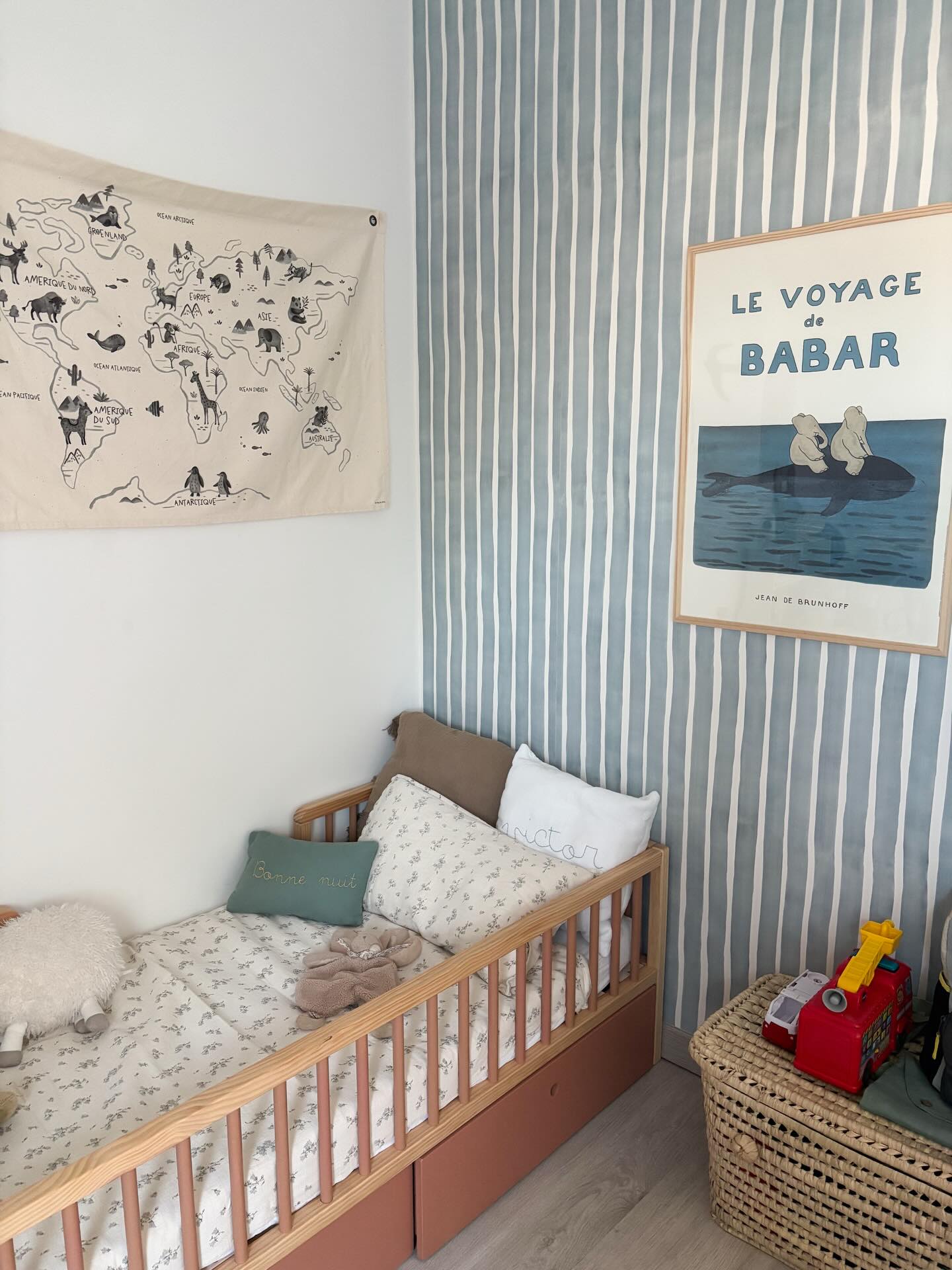 Child's bed with world map tapestry and Le Voyage de Babar poster on striped walls.
