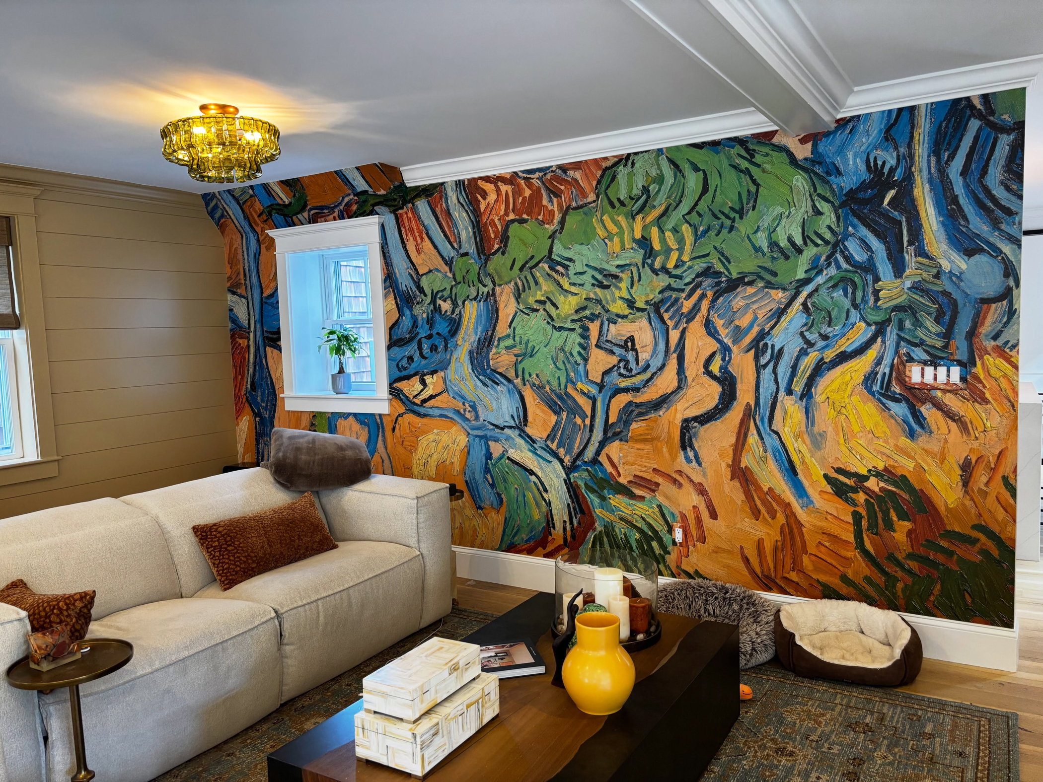 Living room with a vibrant mural of blue and green trees against an orange-yellow background. A light beige sectional sofa is below.