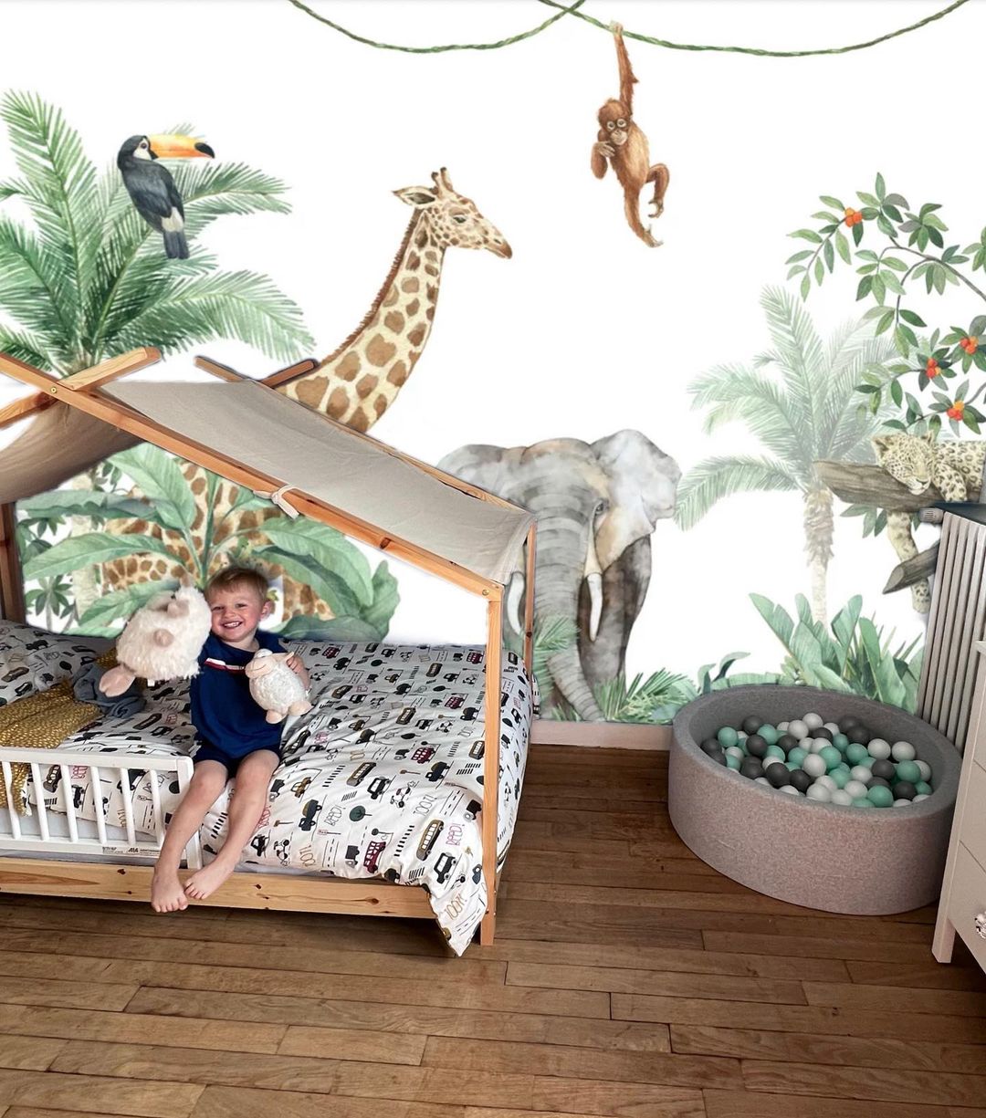Child in a canopy bed with stuffed animals, jungle mural backdrop, and ball pit.