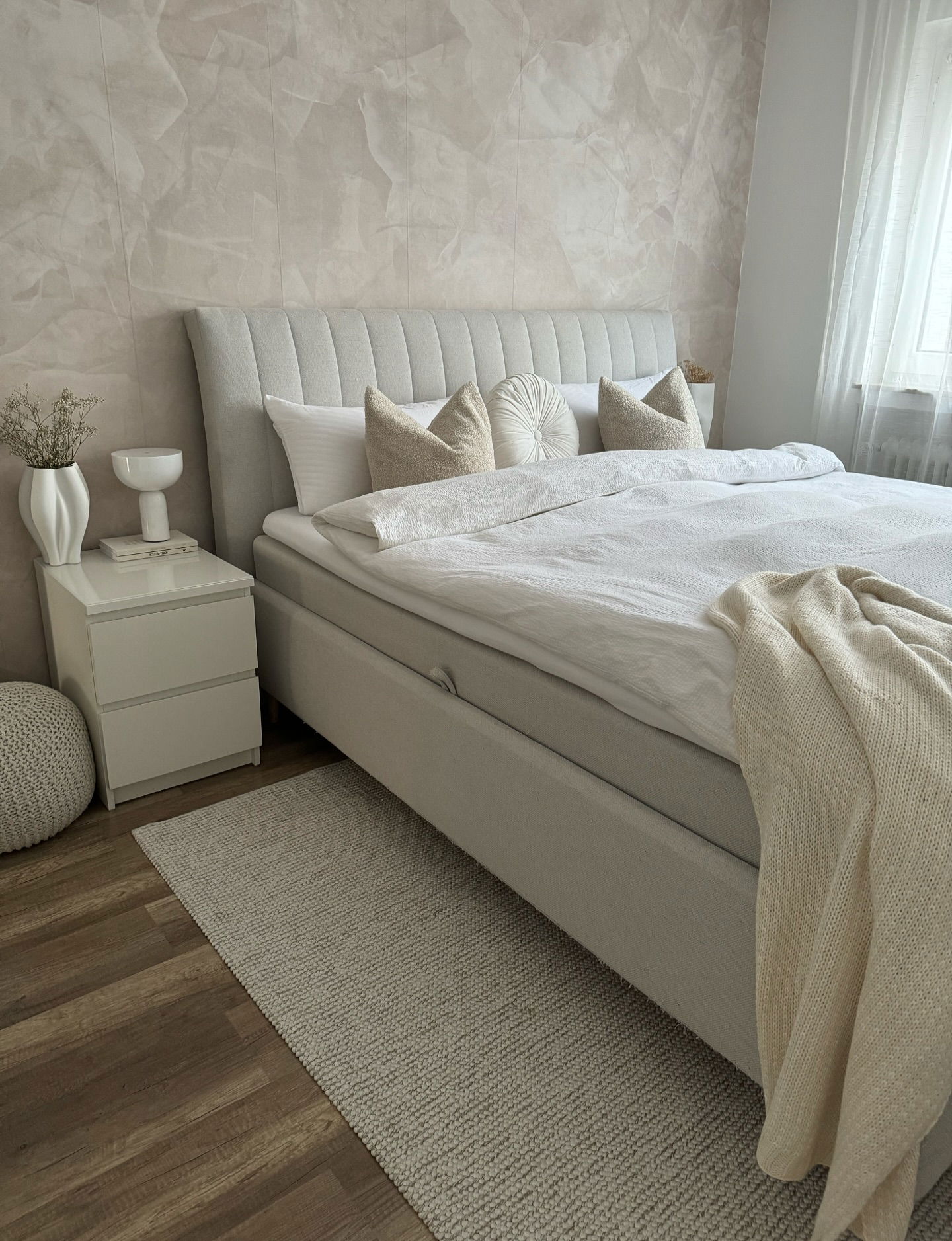 Bedroom with upholstered bed, white linens, and textured wall. Nightstand with vase and bowl. Neutral color scheme.