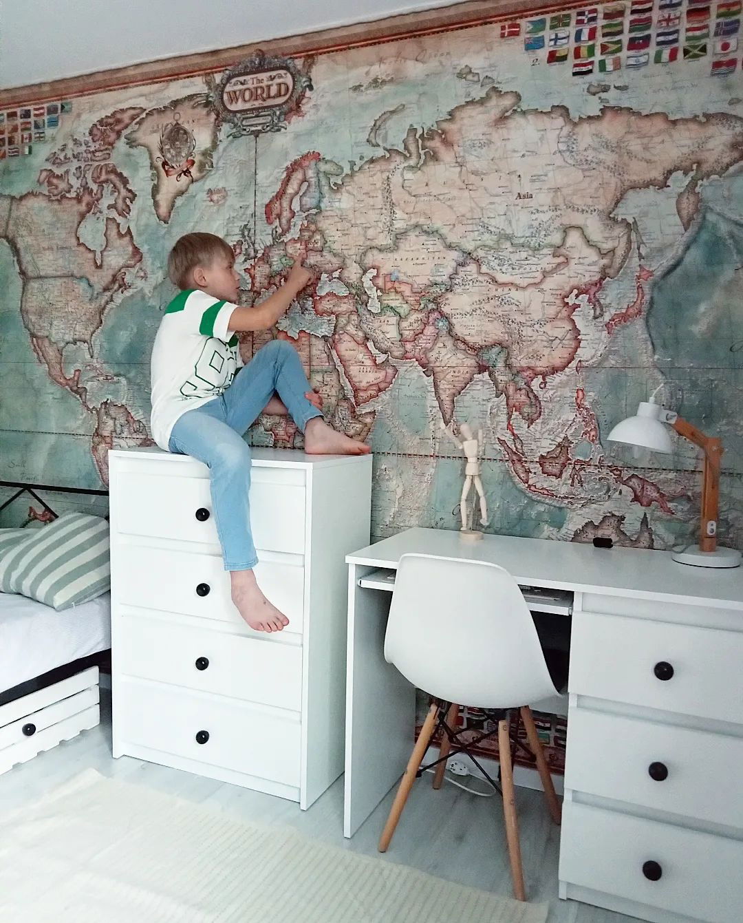 Boy sitting on white dresser, pointing at world map wallpaper in bedroom with desk and chair.