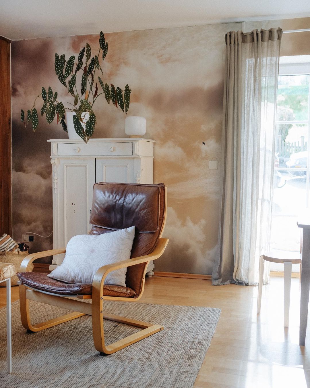 Vue intérieure d'un fauteuil en cuir marron avec un oreiller blanc sur un tapis tissé. Une armoire avec une plante est placée contre un mur à motifs de nuages.