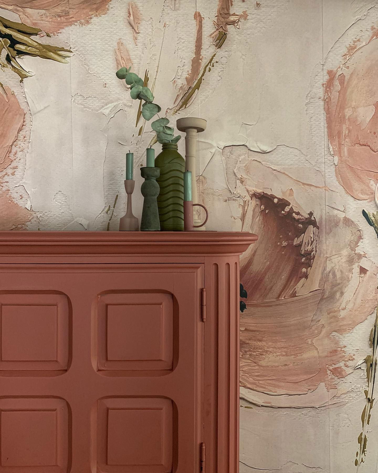 Close-up of a pink cabinet with candles, vase, and floral wallpaper.