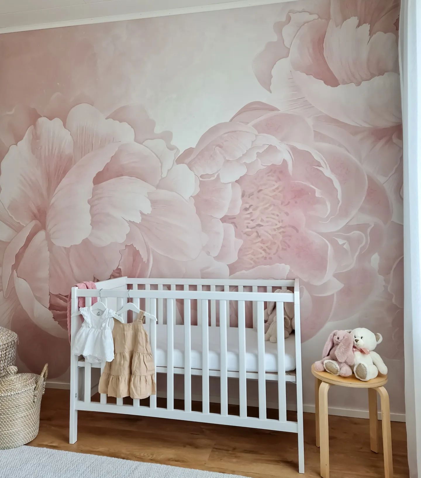 Nursery with white crib against a pink floral mural. Clothes hang on the crib, and stuffed animals sit on a stool.
