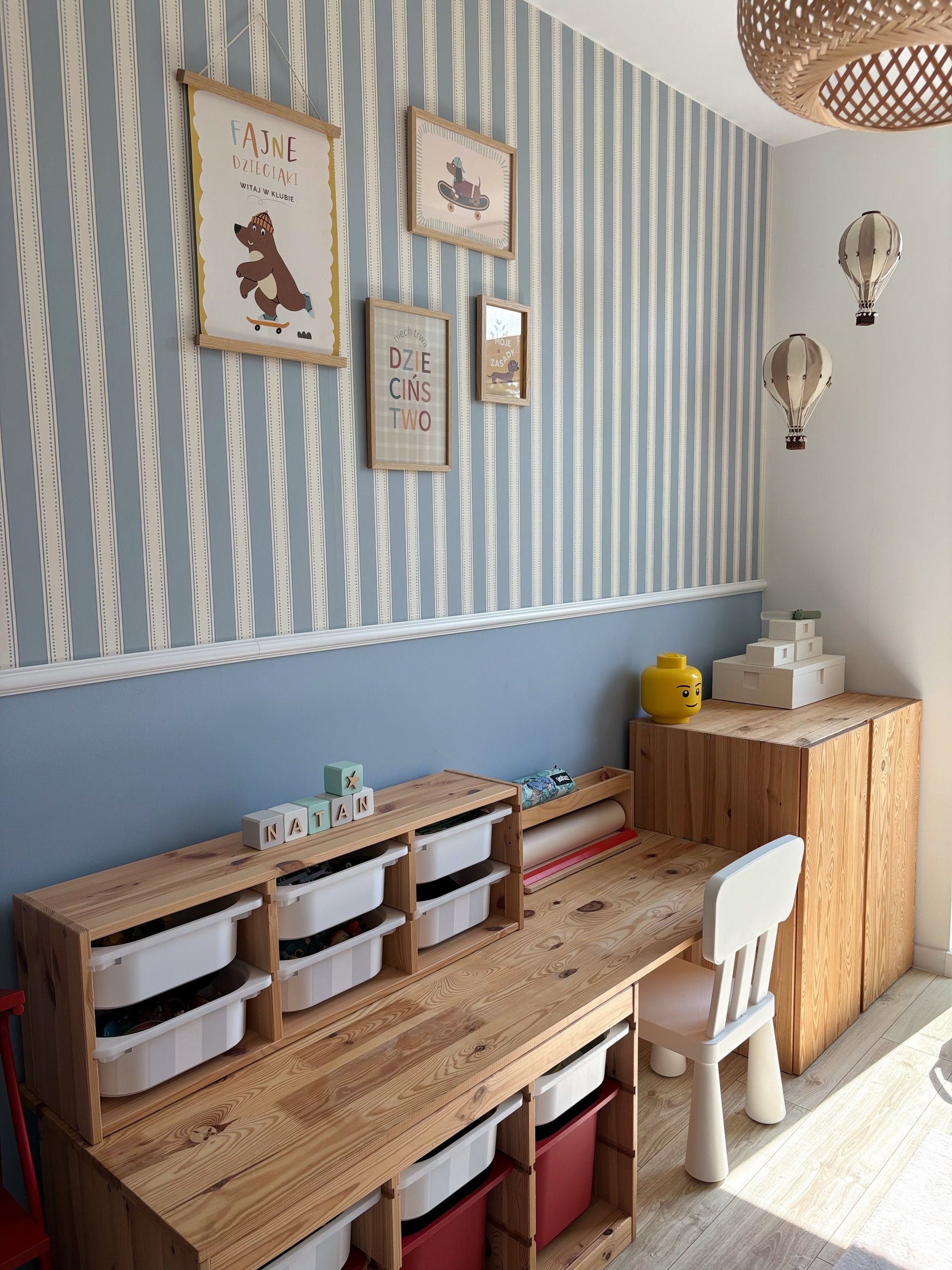 Ein Kinderzimmer mit blau-weiß gestreifter Tapete, einem Schreibtisch aus hellem Holz mit weißen und roten Spielzeugkisten, gerahmten Kunstwerken und dekorativen Heißluftballons.