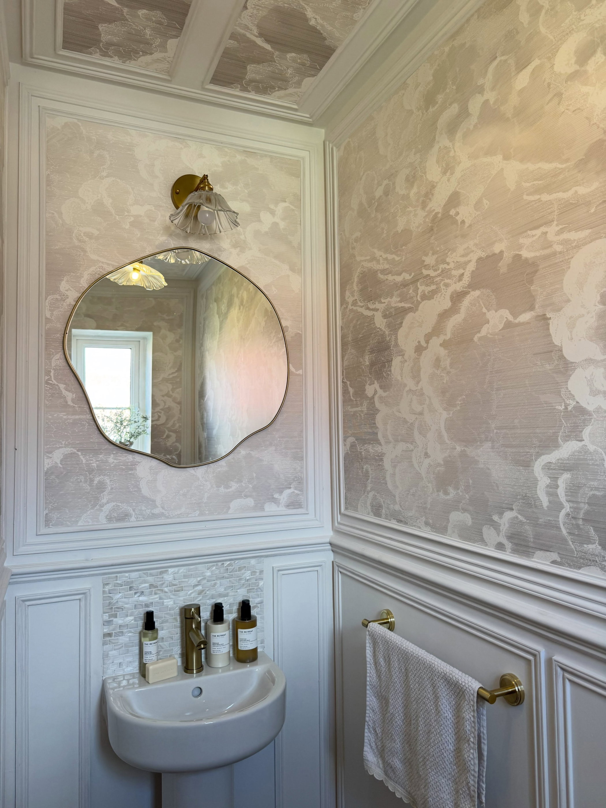 Powder room with light cloud wallpaper, white wainscoting, a gold-framed wavy mirror above a white pedestal sink with gold faucet and toiletries.