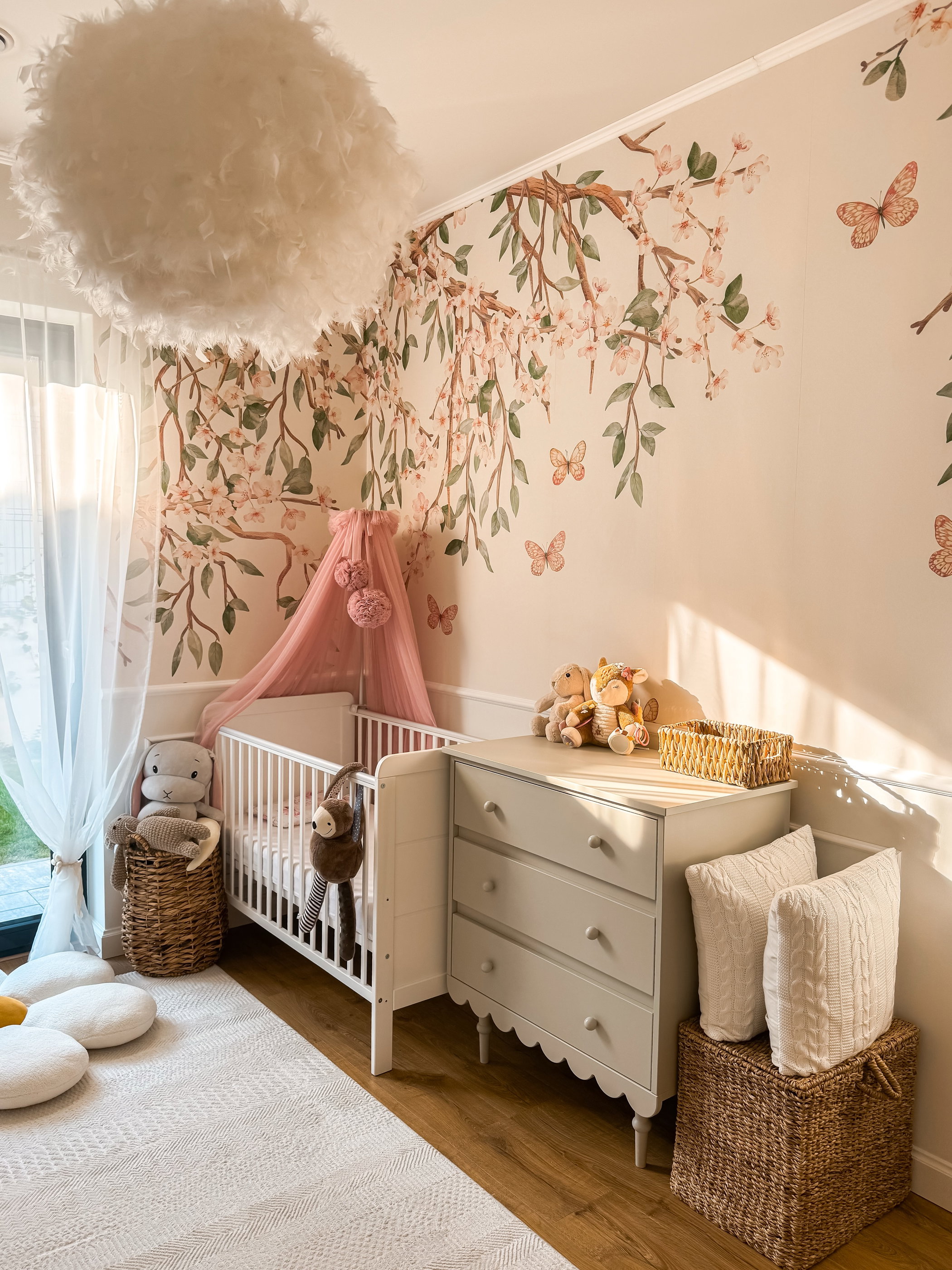 Chambre de bébé avec berceau blanc et baldaquin rose, commode gris clair et lampe de plafond blanche à plumes. Les murs sont décorés de motifs de fleurs de cerisier et de papillons.