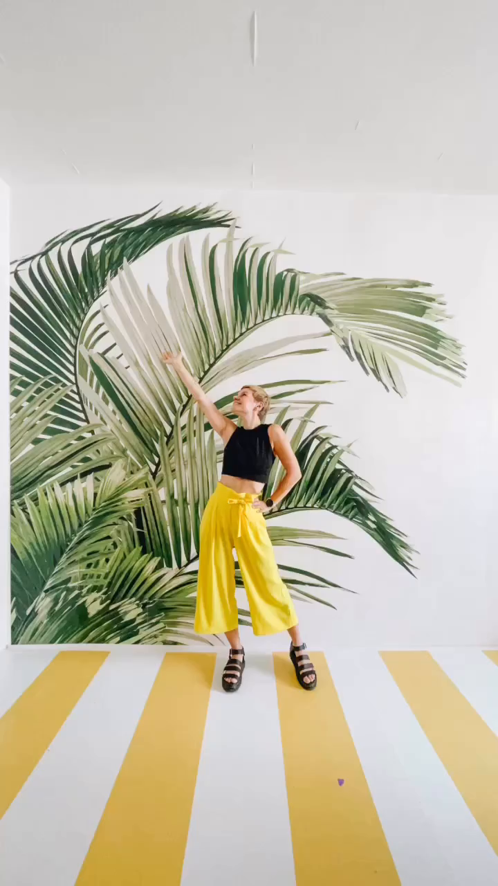 Woman in black crop top and yellow pants poses in front of palm leaf mural.