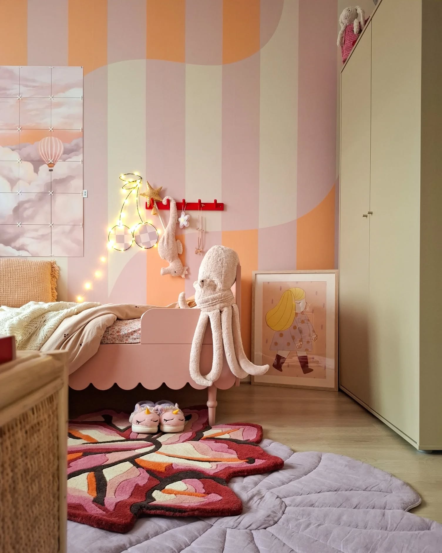 Et barnerom med en rosa, skjellformet seng, et sommerfuglformet teppe foran, og stripete tapet i rosa, oransje og beige. Et veggbilde av en varmluftsballong er til venstre.