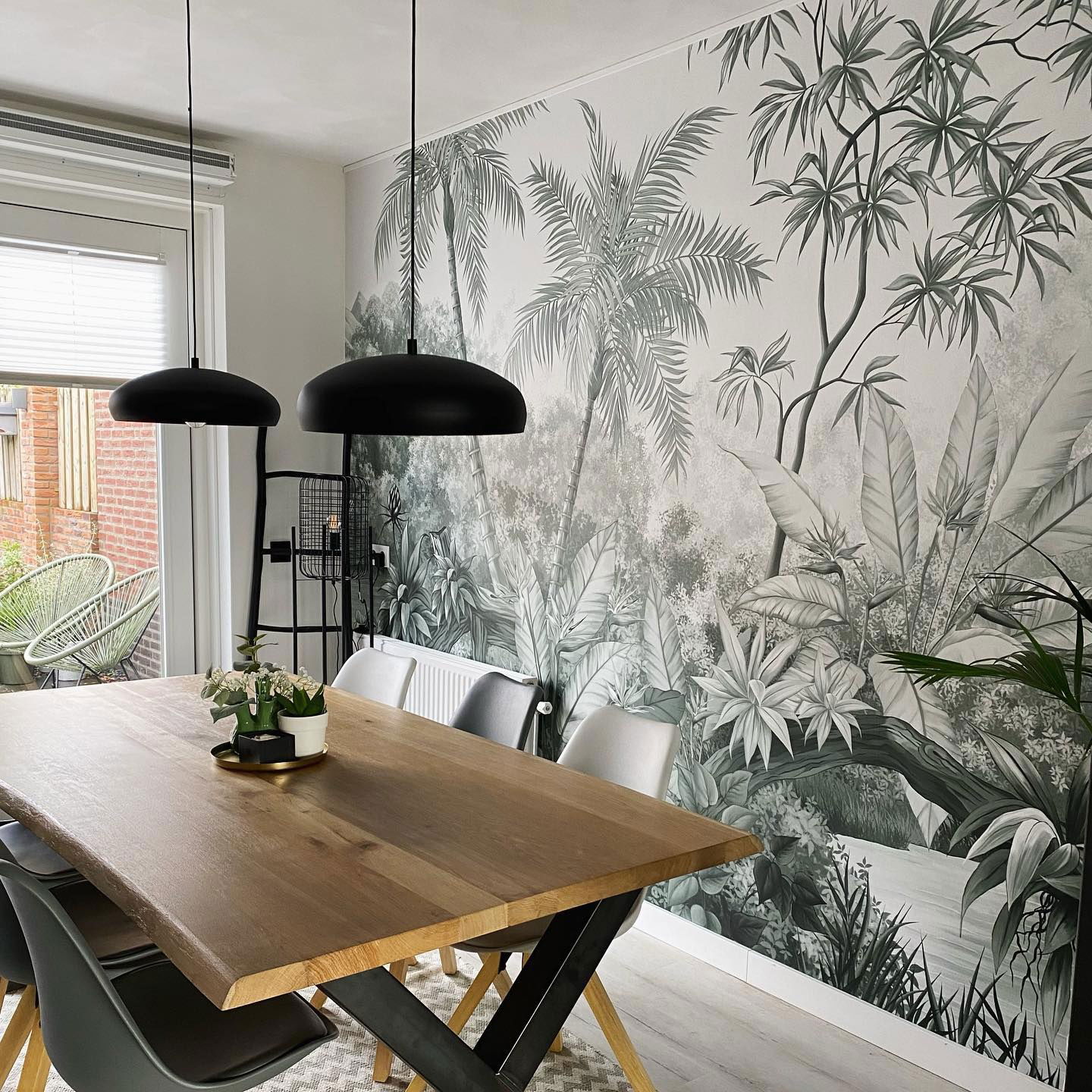 Dining room with wood table, gray chairs, and tropical mural. Black pendant lights hang above.