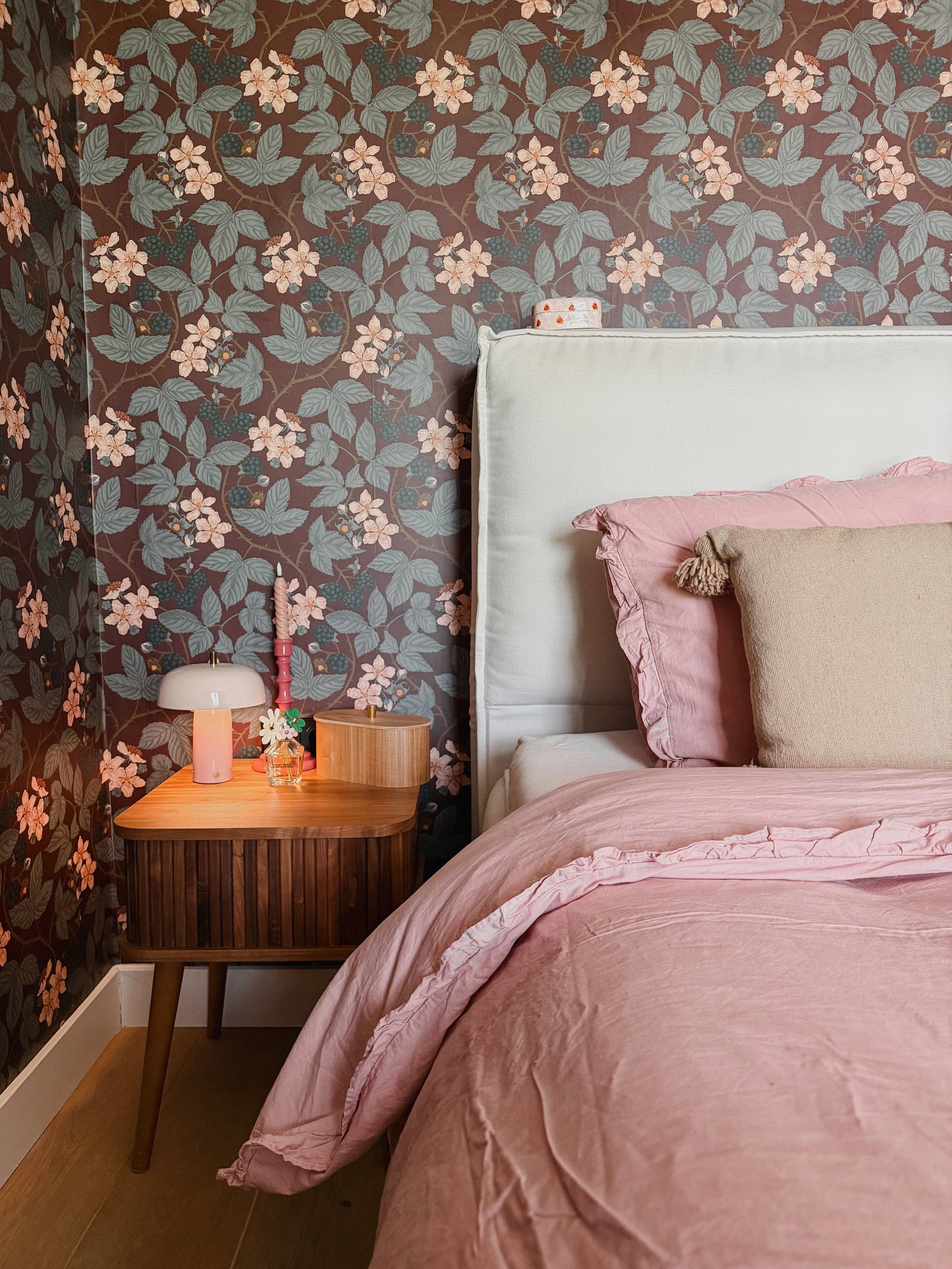 Seng med rosa sengetøy ved siden av et nattbord i tre med en rosa lampe og blomstret tapet