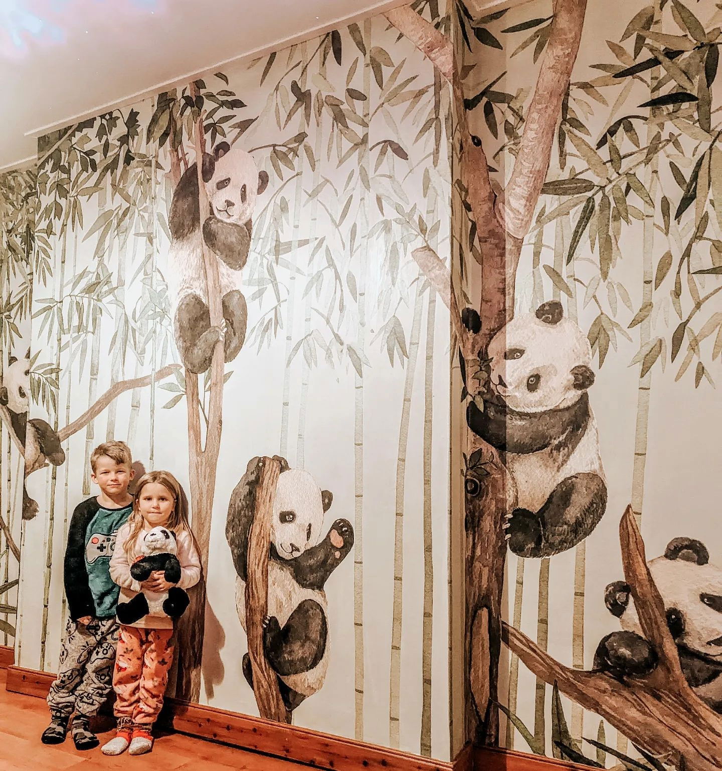 Two children pose in front of a panda and bamboo wallpaper mural.