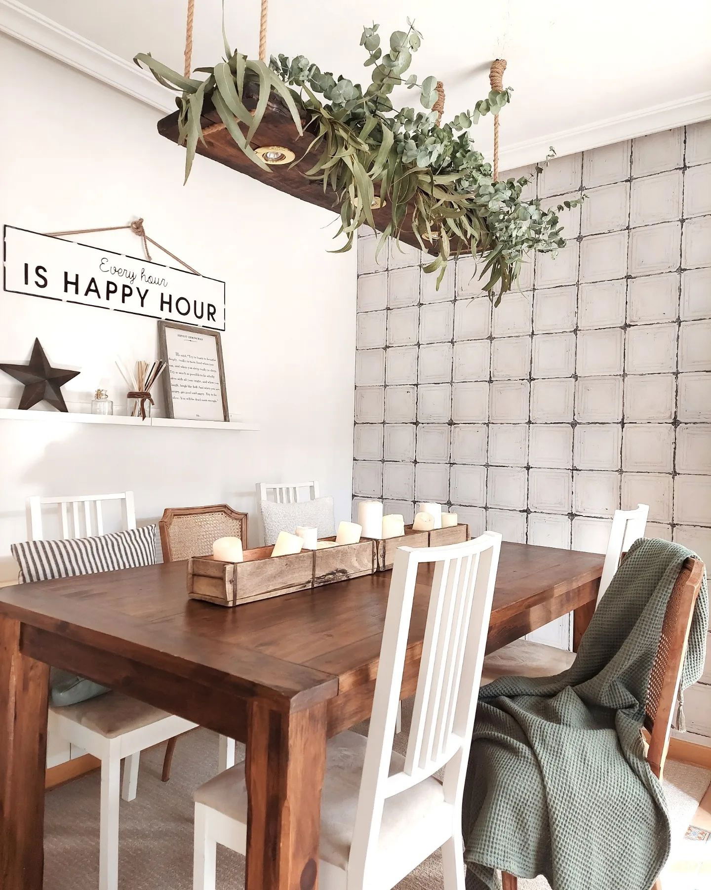 Esszimmer mit Holztisch, weißen Stühlen und hängendem Eukalyptus-Mittelstück. Schild "Every Hour is Happy Hour" an der Wand.