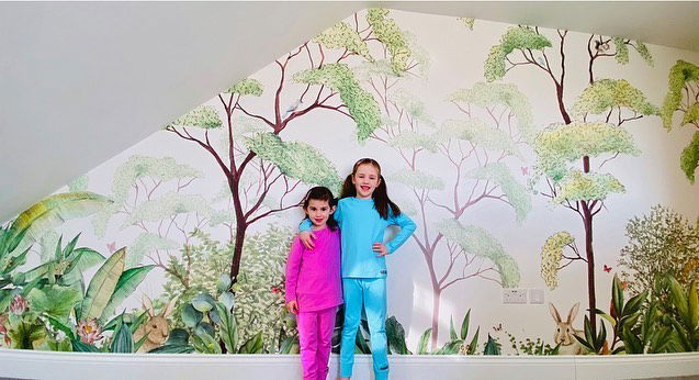 Dos chicas en pijama están de pie frente a un mural. El mural representa una exuberante escena de bosque con árboles, plantas y conejos.