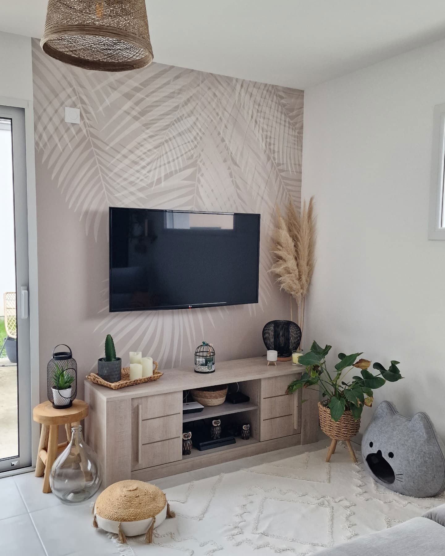 Sala de estar con televisor sobre papel tapiz de hojas de palma, mesa consola de madera clara y decoración tejida.