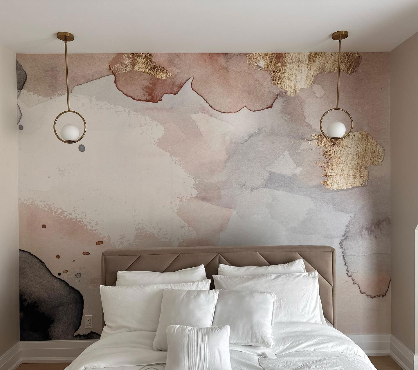 Bedroom with white bedding, tan headboard, and watercolor-style wallpaper with gold accents. Two gold pendant lights hang above.