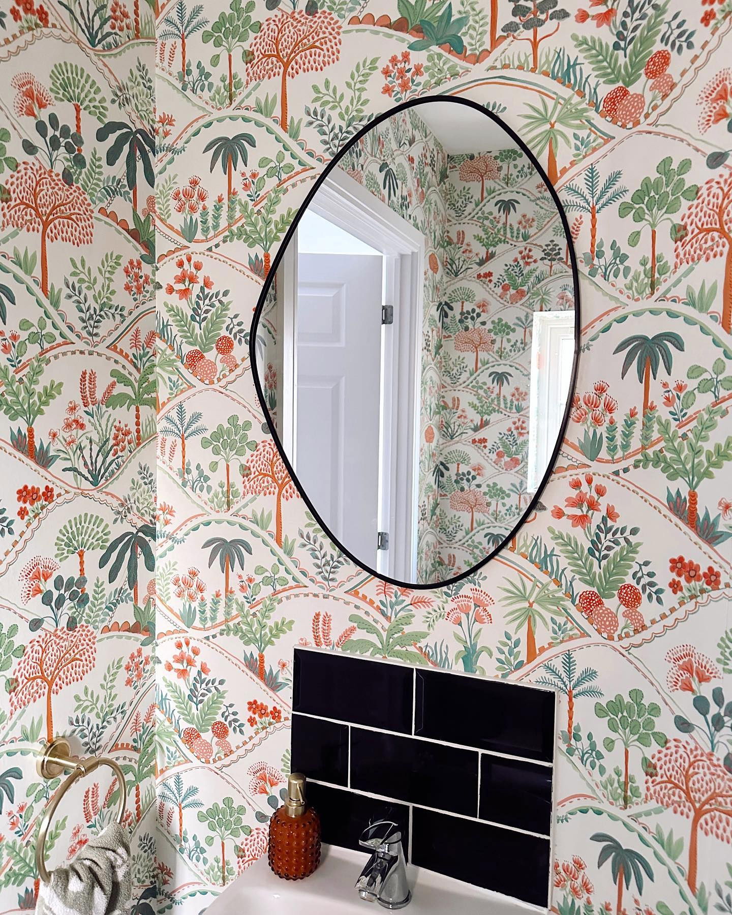 Badkamer met bloemenbehang, zwarte metrotegels en ovale spiegel.
