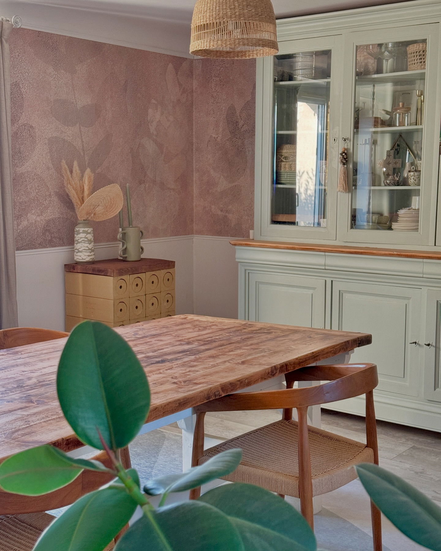 Salle à manger avec table en bois, chaises et buffet vert clair. Papier peint avec motif de feuilles. Suspension en osier.