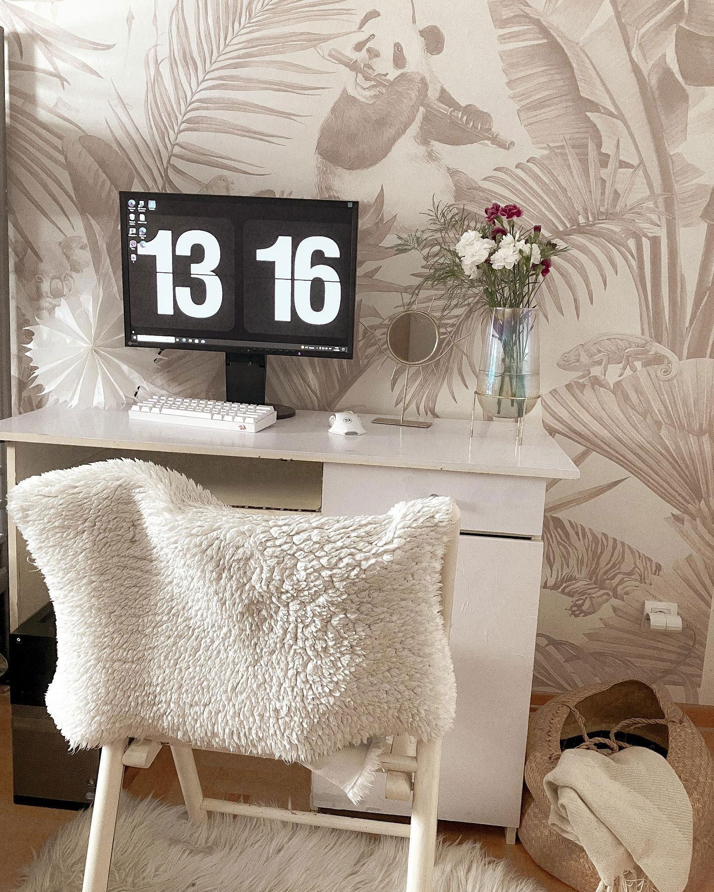 Installation de bureau avec un ordinateur affichant «&nbsp;13&nbsp;16&nbsp;», des fleurs et une chaise moelleuse, sur un papier peint tropical.