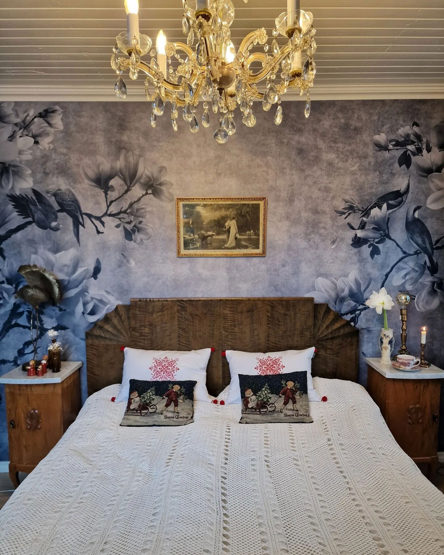 Schlafzimmer mit floraler Tapete, hölzernem Kopfteil und weißer Häkeldecke. Ein Kristalllüster hängt darüber.