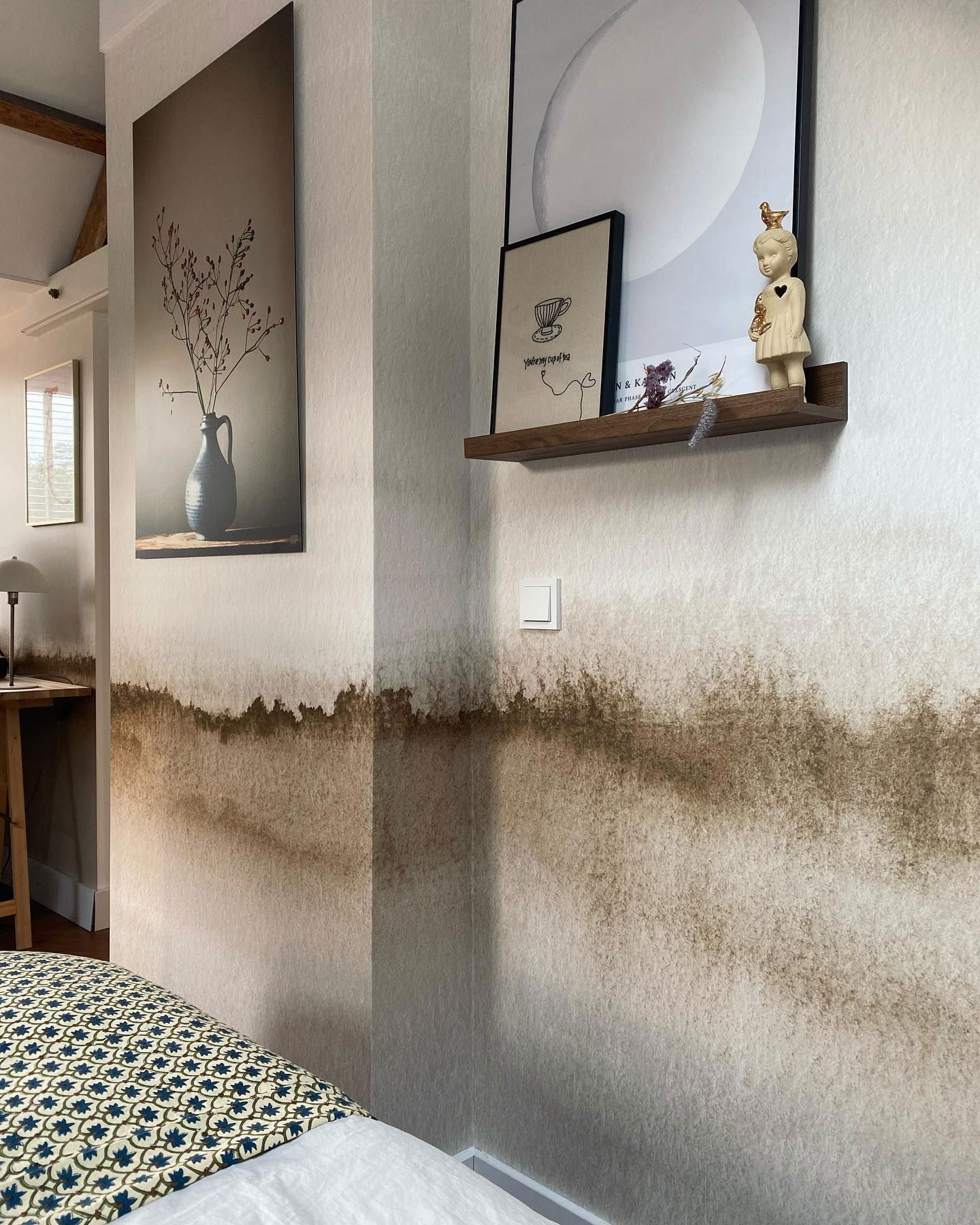Bedroom corner with brown ombre wallpaper, shelf with art prints, and vase painting.
