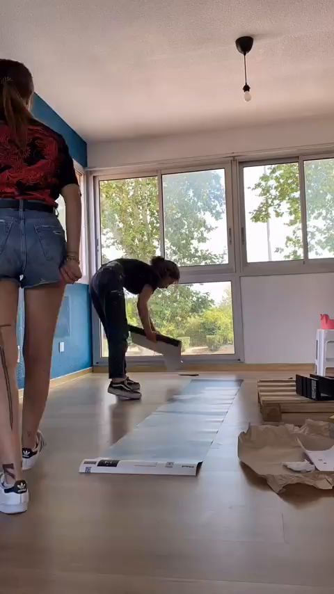 Two women lay out flooring in a room with large windows.