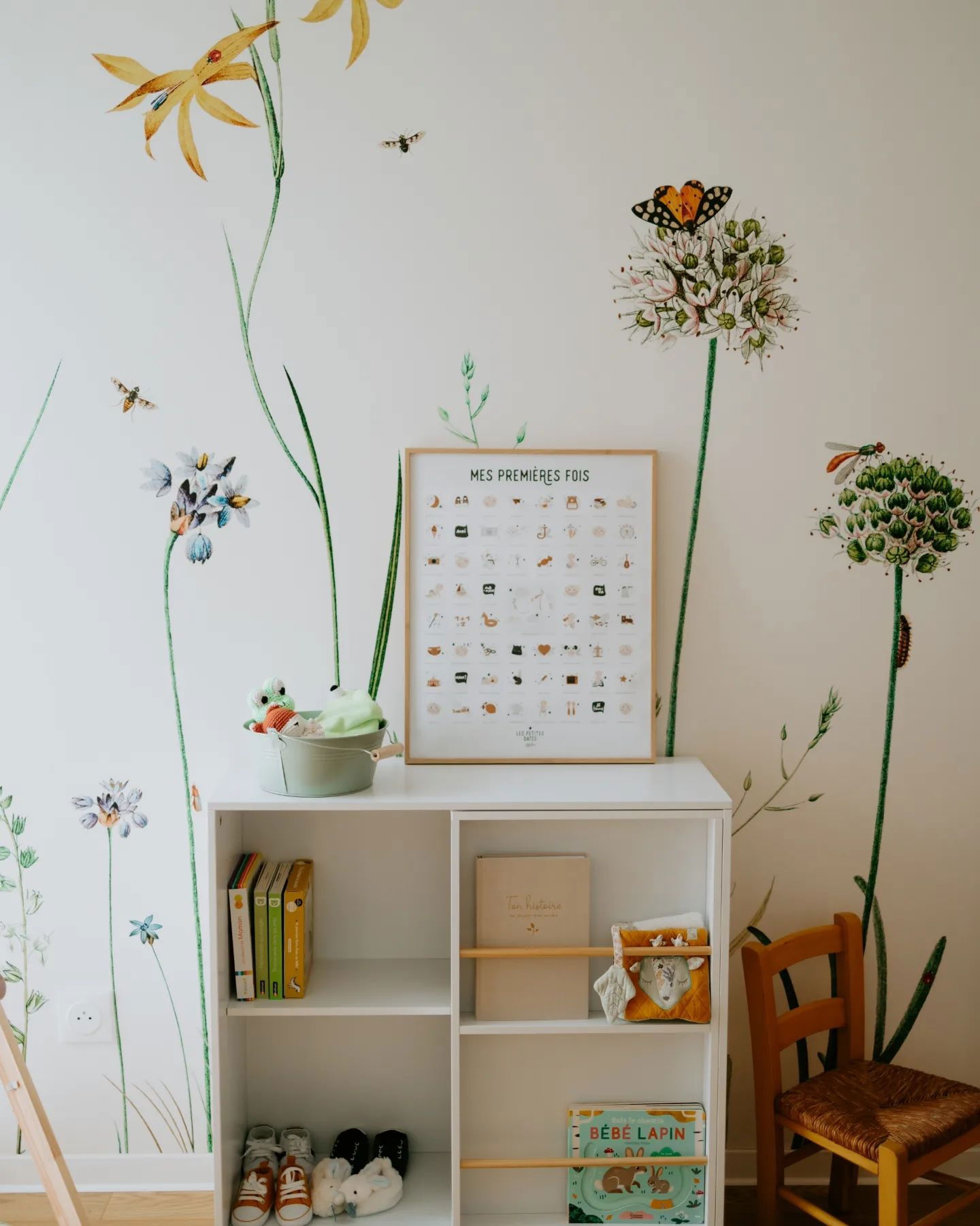 Chambre d'enfant avec des autocollants muraux floraux, une étagère blanche, des livres, des jouets et une affiche encadrée "Mes Premieres Fois".