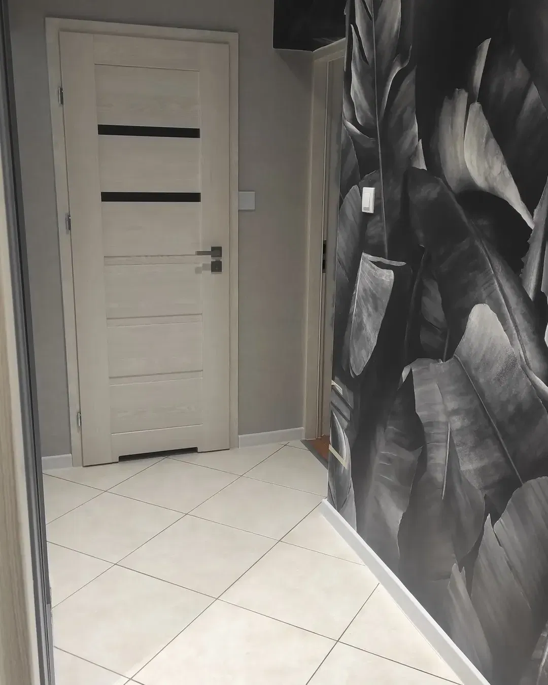 Couloir avec porte blanche, carrelage et papier peint à feuilles noir et blanc.