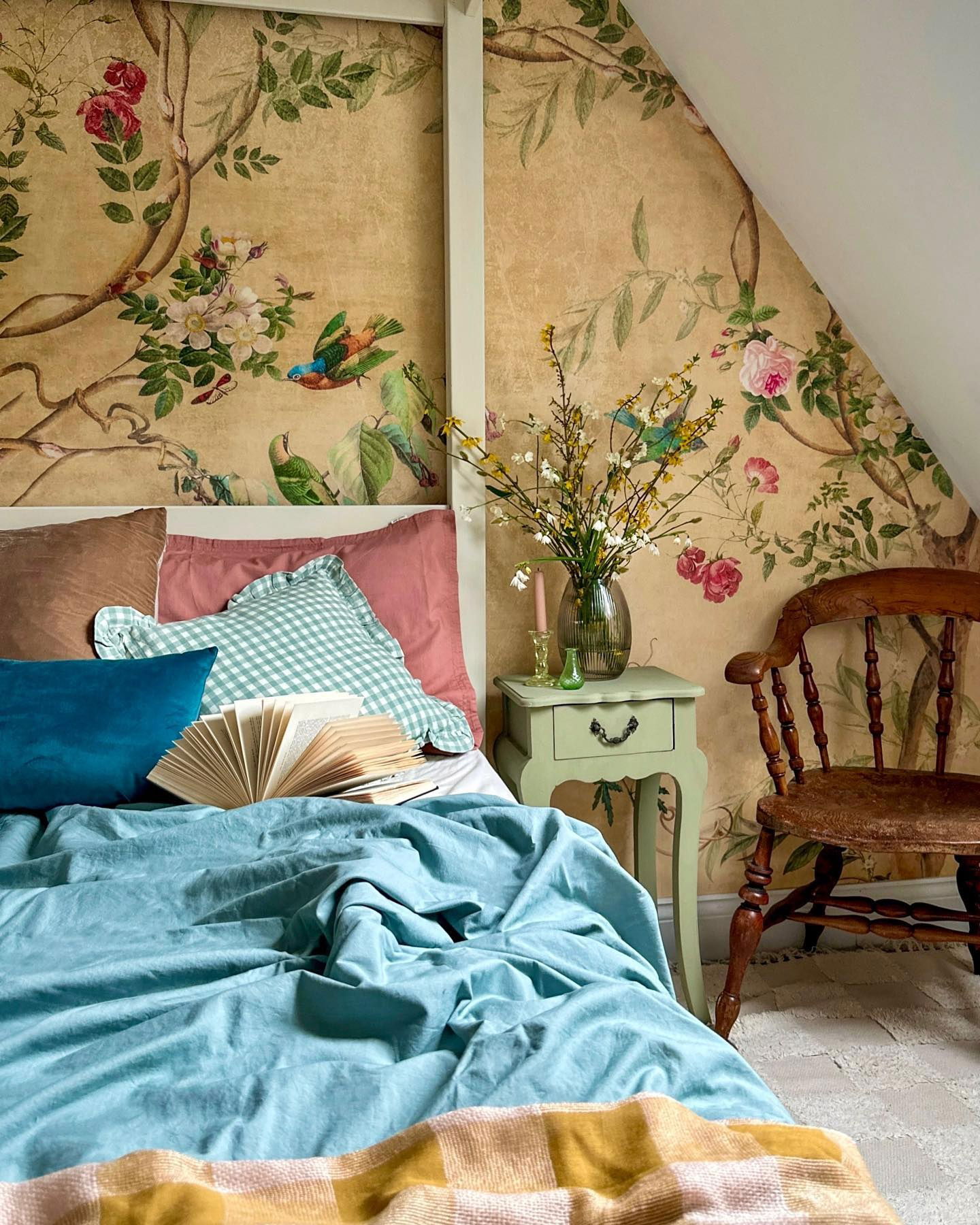 Bedroom with floral wallpaper, blue bedding, and a vintage wooden chair. Nightstand with flowers.