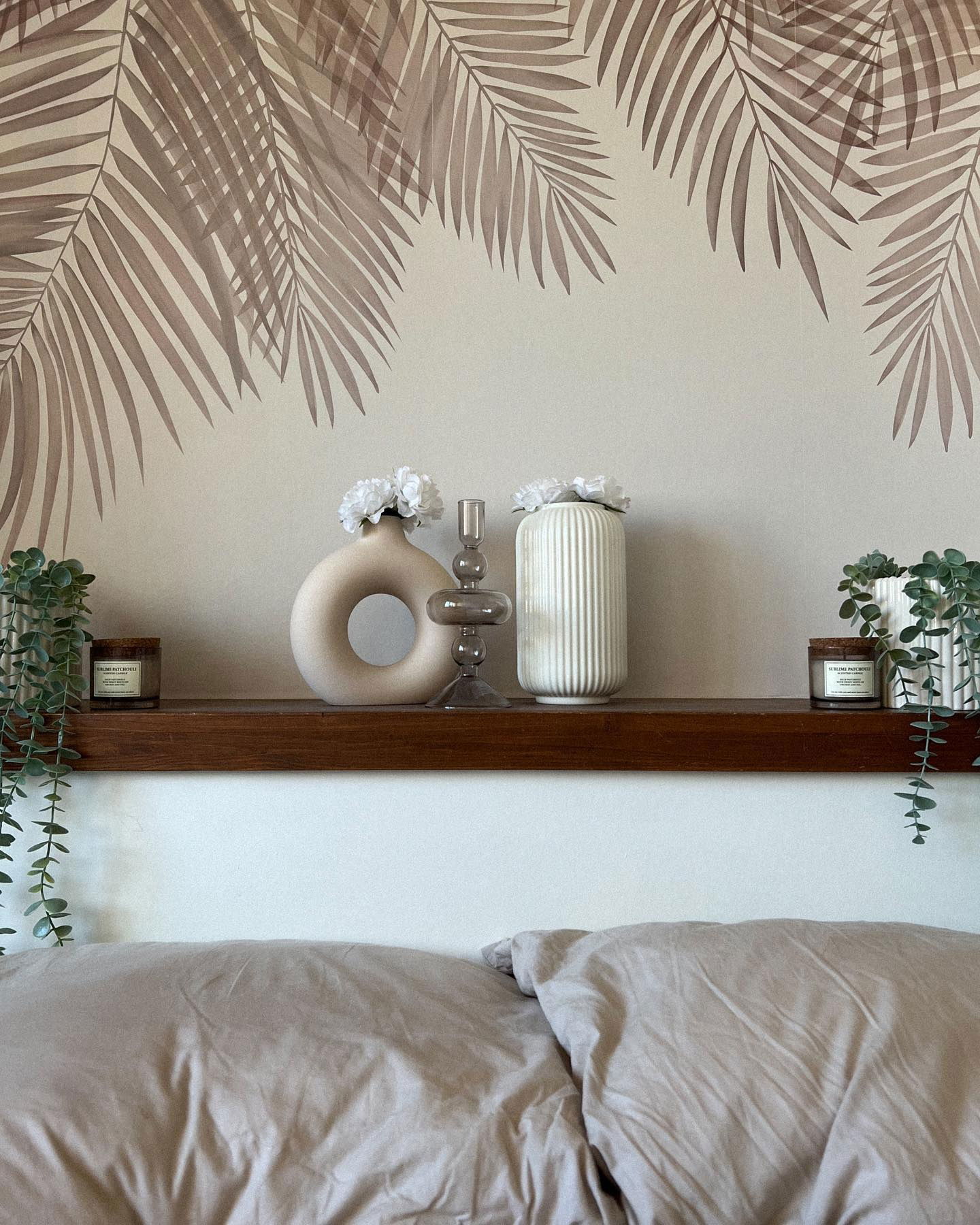 Decoración de dormitorio con arte de pared de hojas de palma, jarrones, vela y plantas colgantes en un estante encima de una cama.