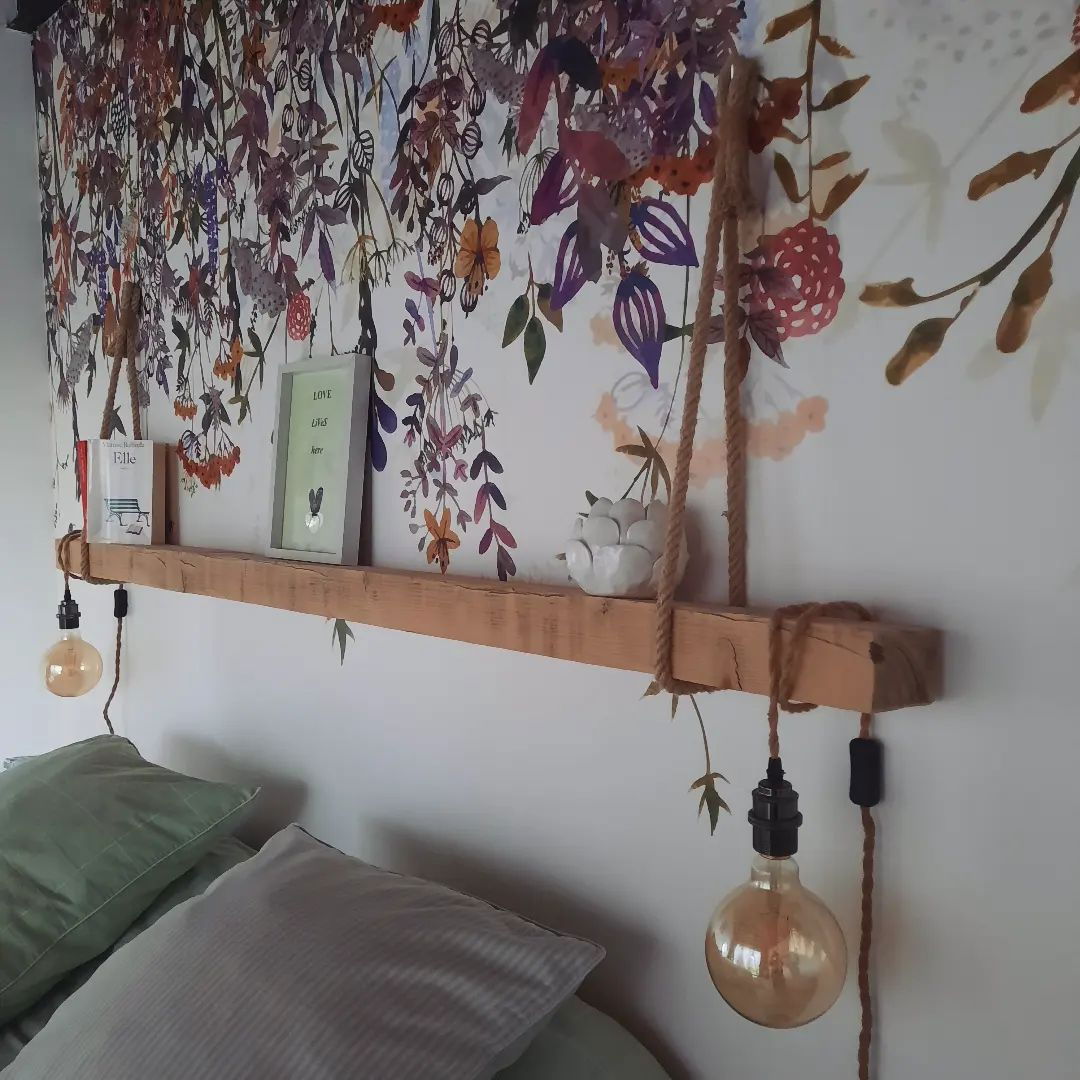 Schlafzimmer mit einem hölzernen Regal-Kopfteil, das an einem Seil hängt, Blumentapete und freiliegender Glühbirnenbeleuchtung.