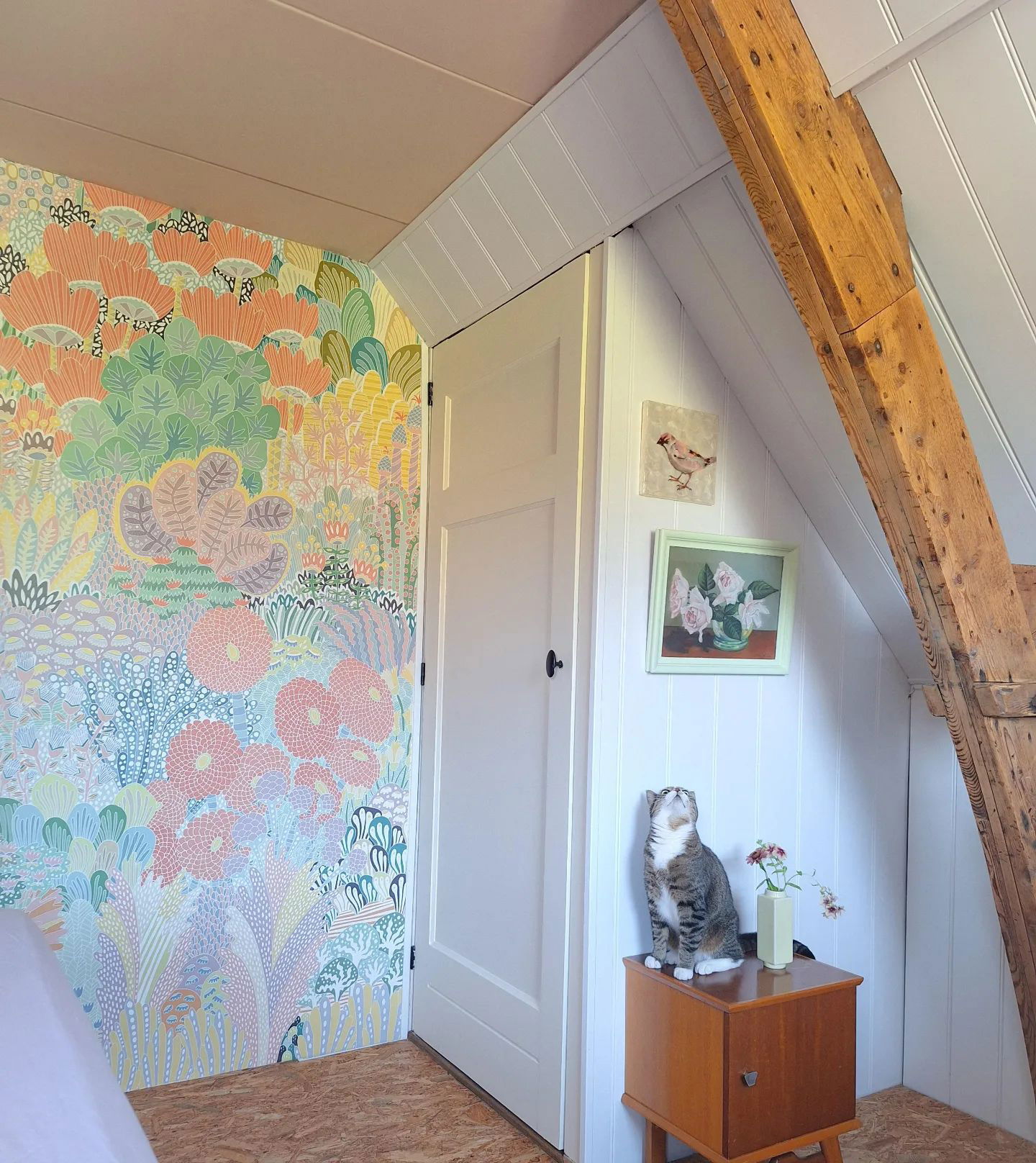 Attic room with floral wallpaper, white door, and tabby cat sitting on a wooden side table.