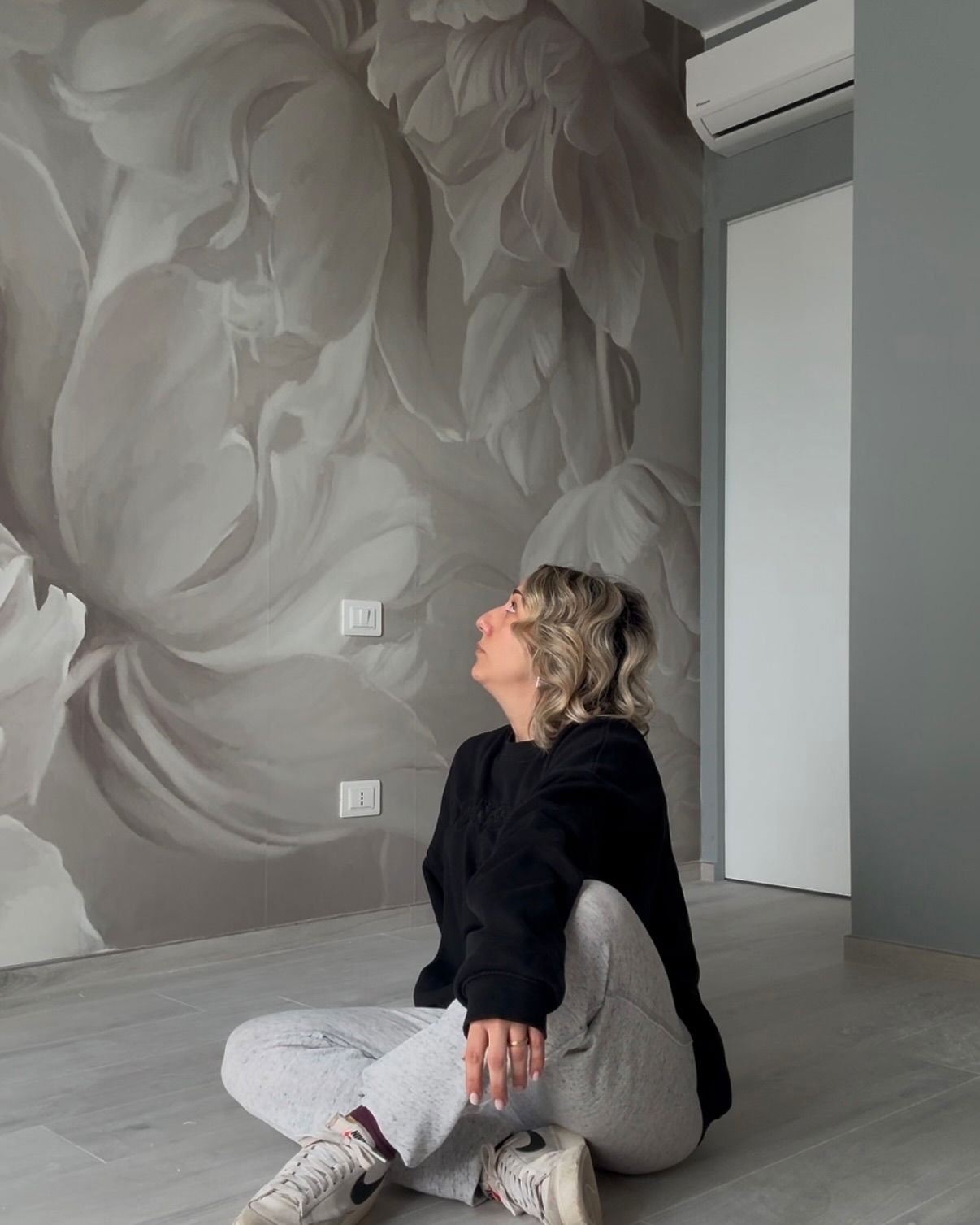 Woman sits cross-legged in front of a wall mural of large, gray flowers.