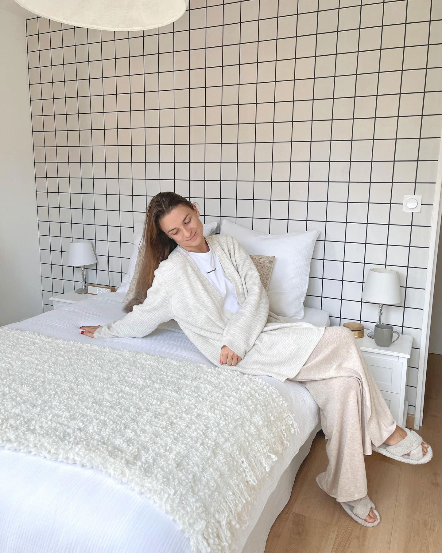Woman in pajamas sitting on bed with grid-patterned wall. She wears a cardigan and fuzzy slippers.