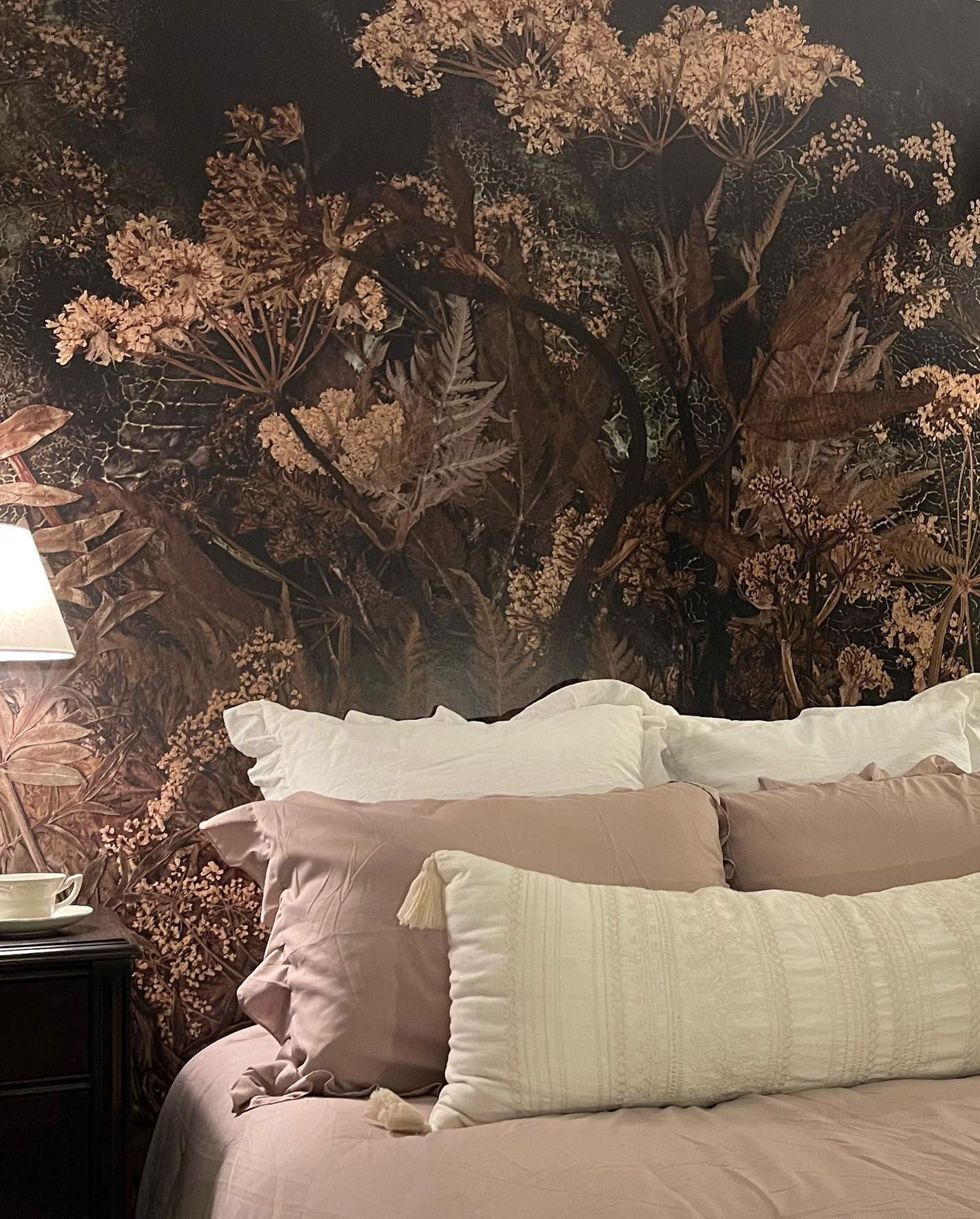 Schlafzimmer mit floraler Tapete, weißer und lilafarbener Bettwäsche und einem dunklen Holz Nachttisch.
