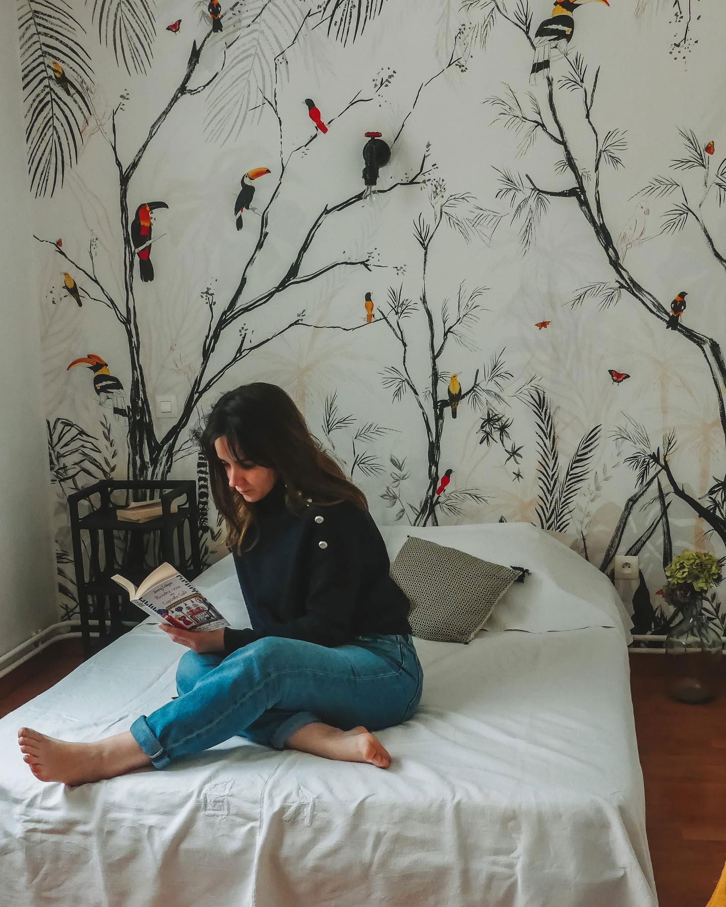 A woman reading a book on a bed with a tropical bird and tree wallpaper.