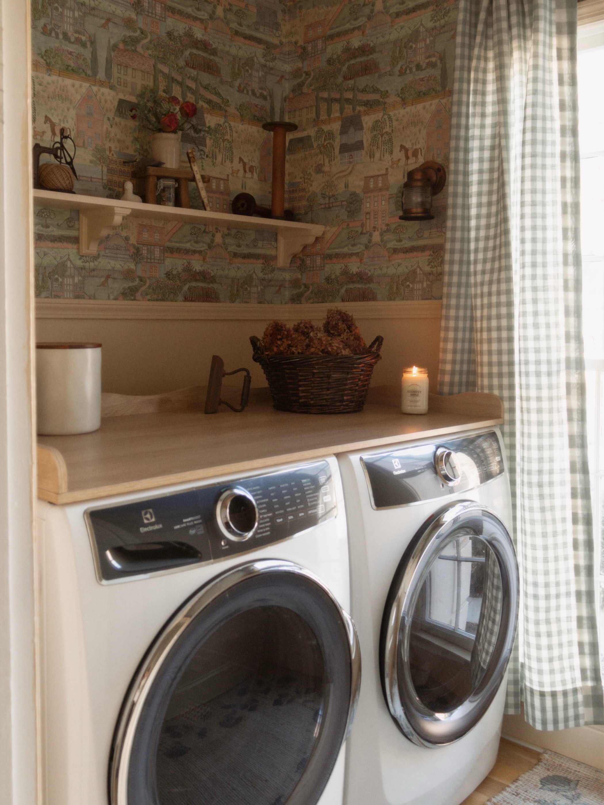 A rustic laundry room featuring white front-load washer and dryer under a wooden counter. The walls have patterned wallpaper and a white shelf with decor. A window with green gingham curtains is on the right.