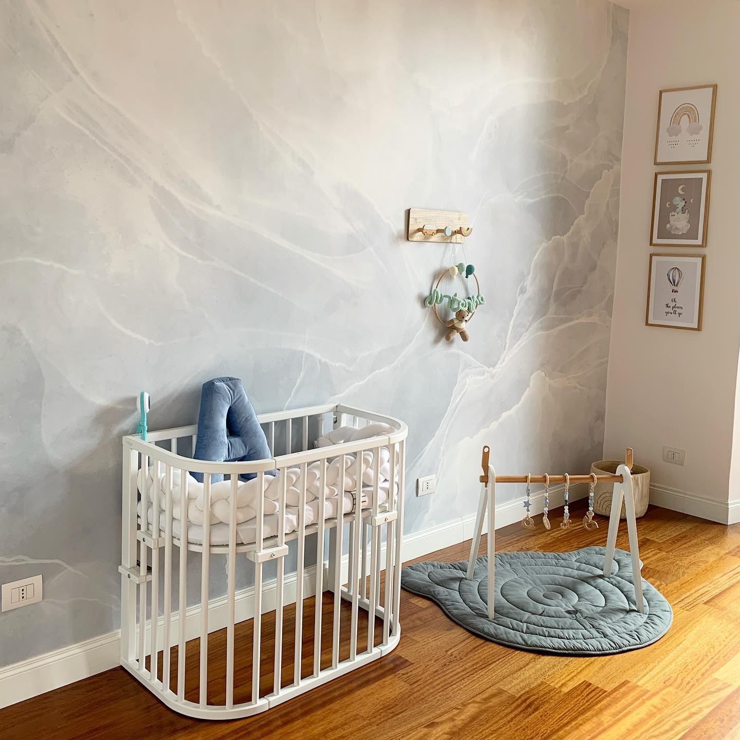 Une chambre de bébé avec un berceau ovale blanc, un tapis de jeu bleu en forme de baleine avec un parc en bois et un mur gris clair à motif marbre.