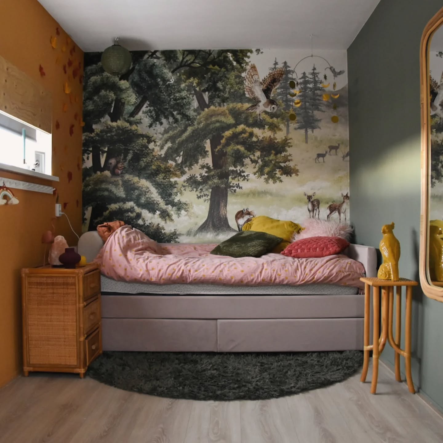 Une chambre avec une fresque forestière représentant des cerfs, un hibou et un renard. Un canapé-lit avec une literie rose, une table de chevet en osier et une table d'appoint.