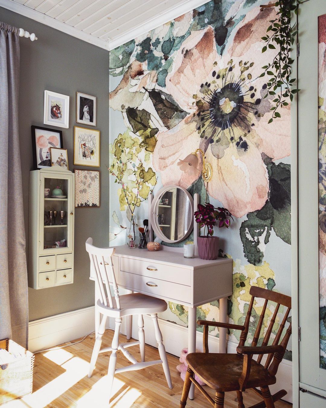 Gemütliches Zimmer mit floraler Tapete, Schminktisch und Stühlen. Wandkunst und Hängeschrank sorgen für Charme. Sonnenlicht auf Holzboden.