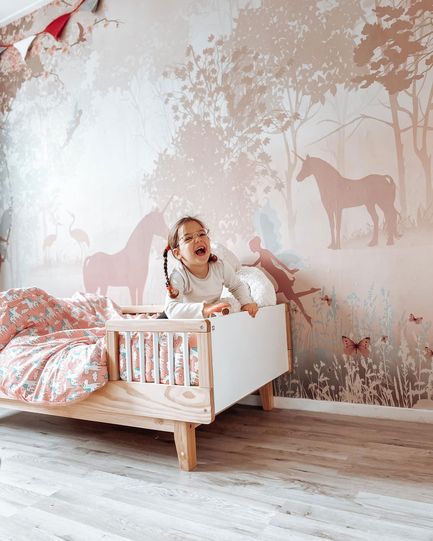 A laughing child with pigtails stands in a wooden toddler bed with unicorn-themed bedding. A unicorn and fairy mural is on the wall.