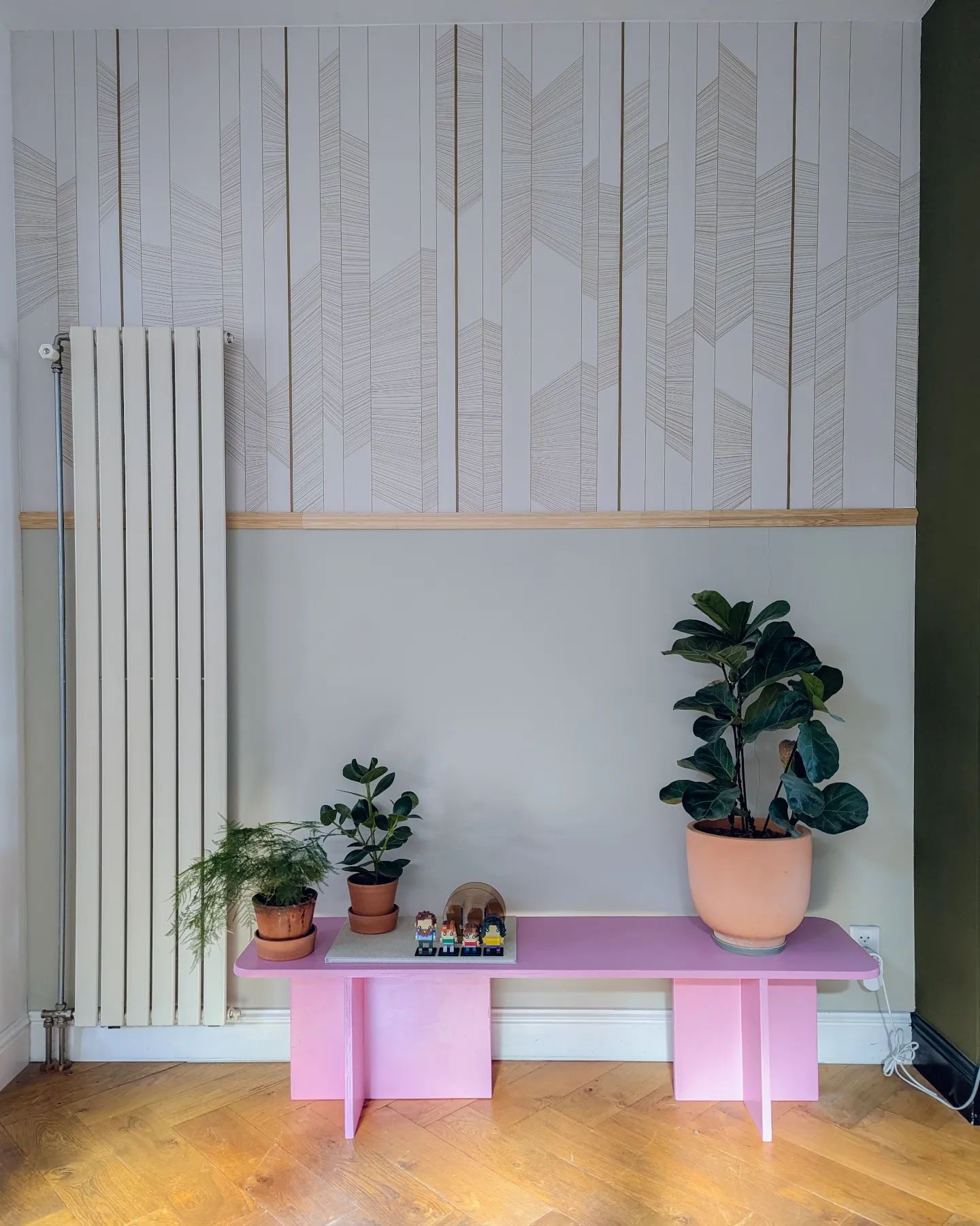 Vista interior con banco rosa que sostiene plantas, contra papel tapiz estampado y pared de dos tonos.